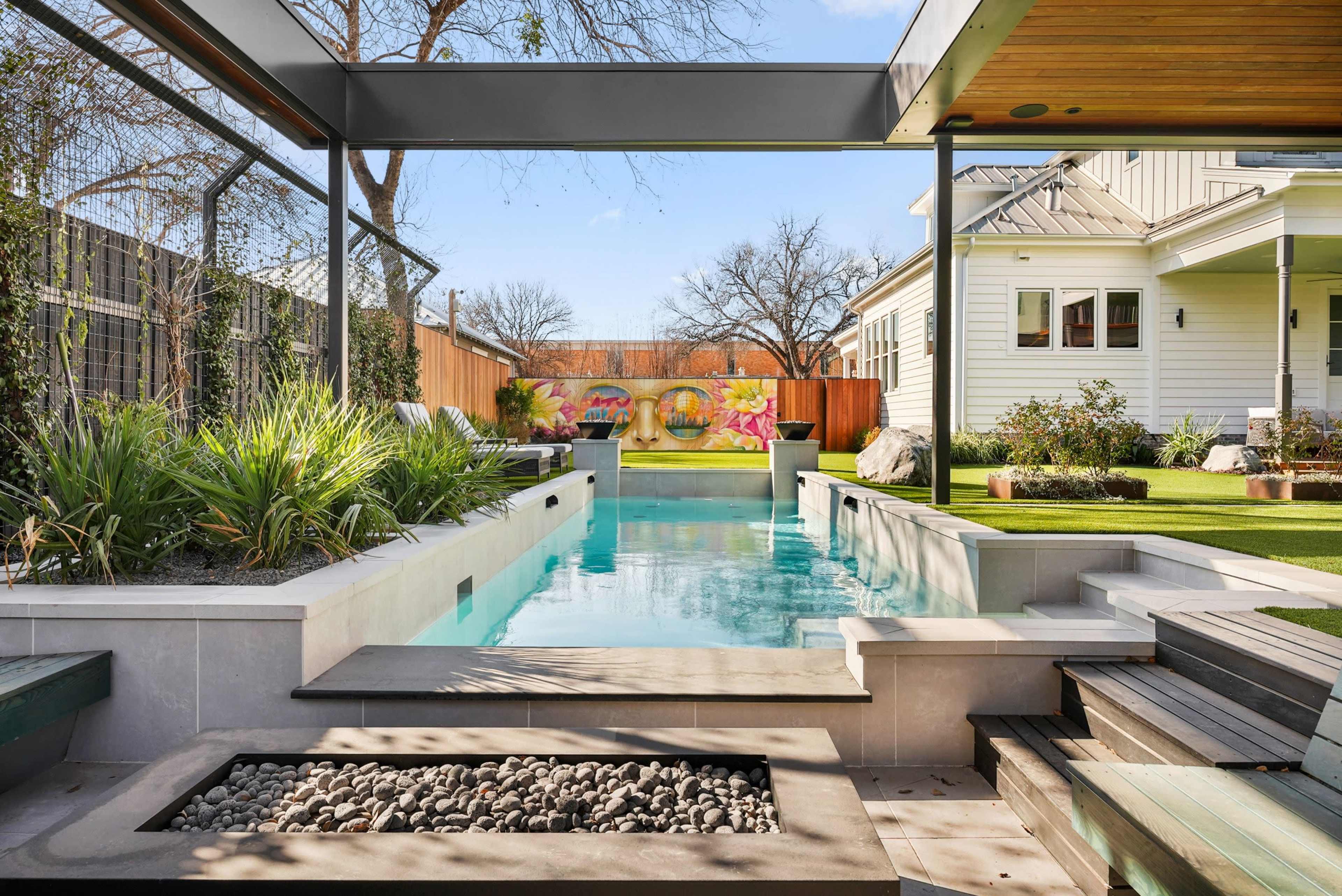 The image shows a modern backyard featuring a rectangular swimming pool surrounded by landscaping and a wooden deck beside a home.