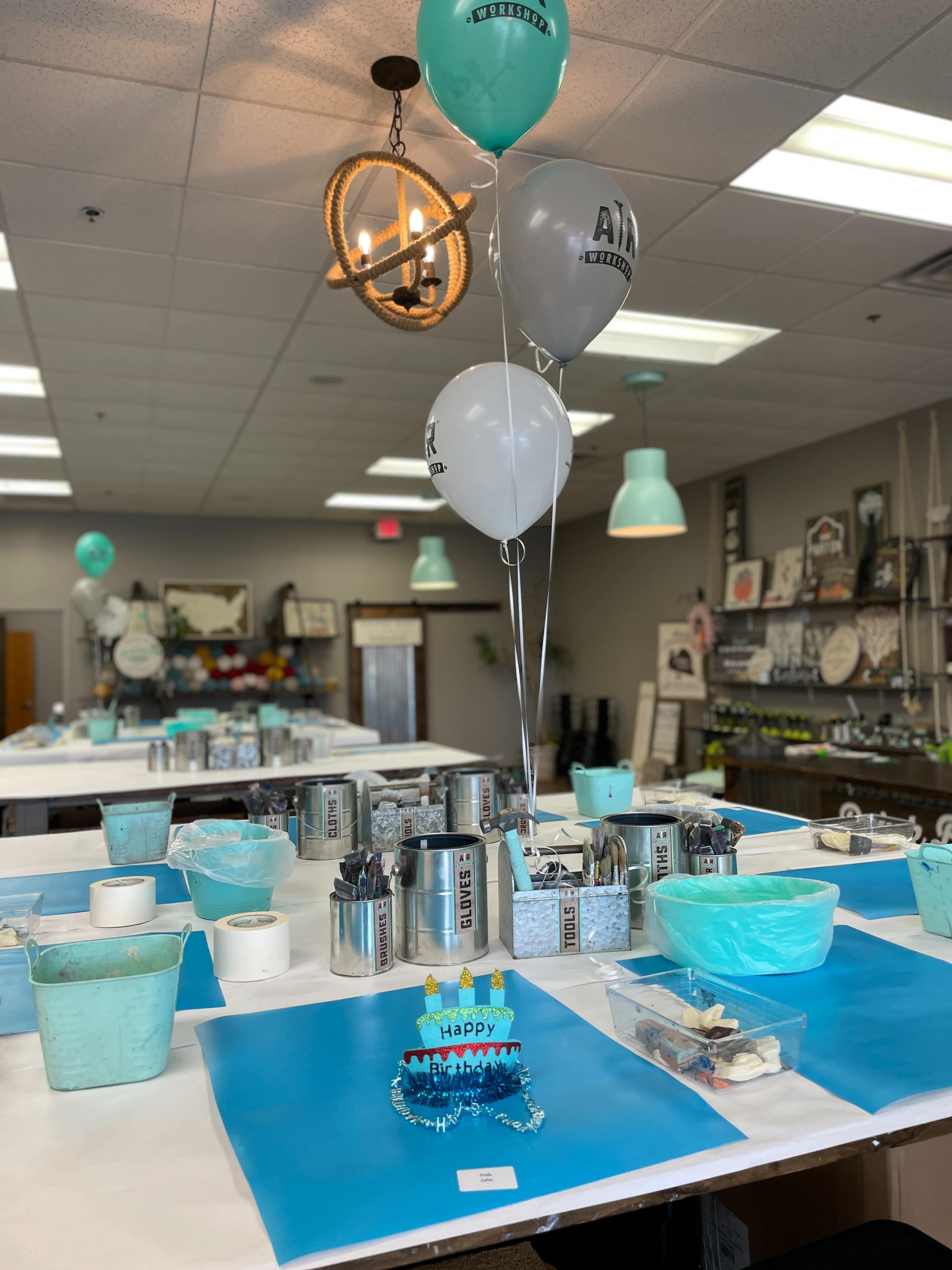 The image shows a creatively arranged workspace with tables covered in blue tablecloths, decorated with balloons and organized supplies for a craft activity.