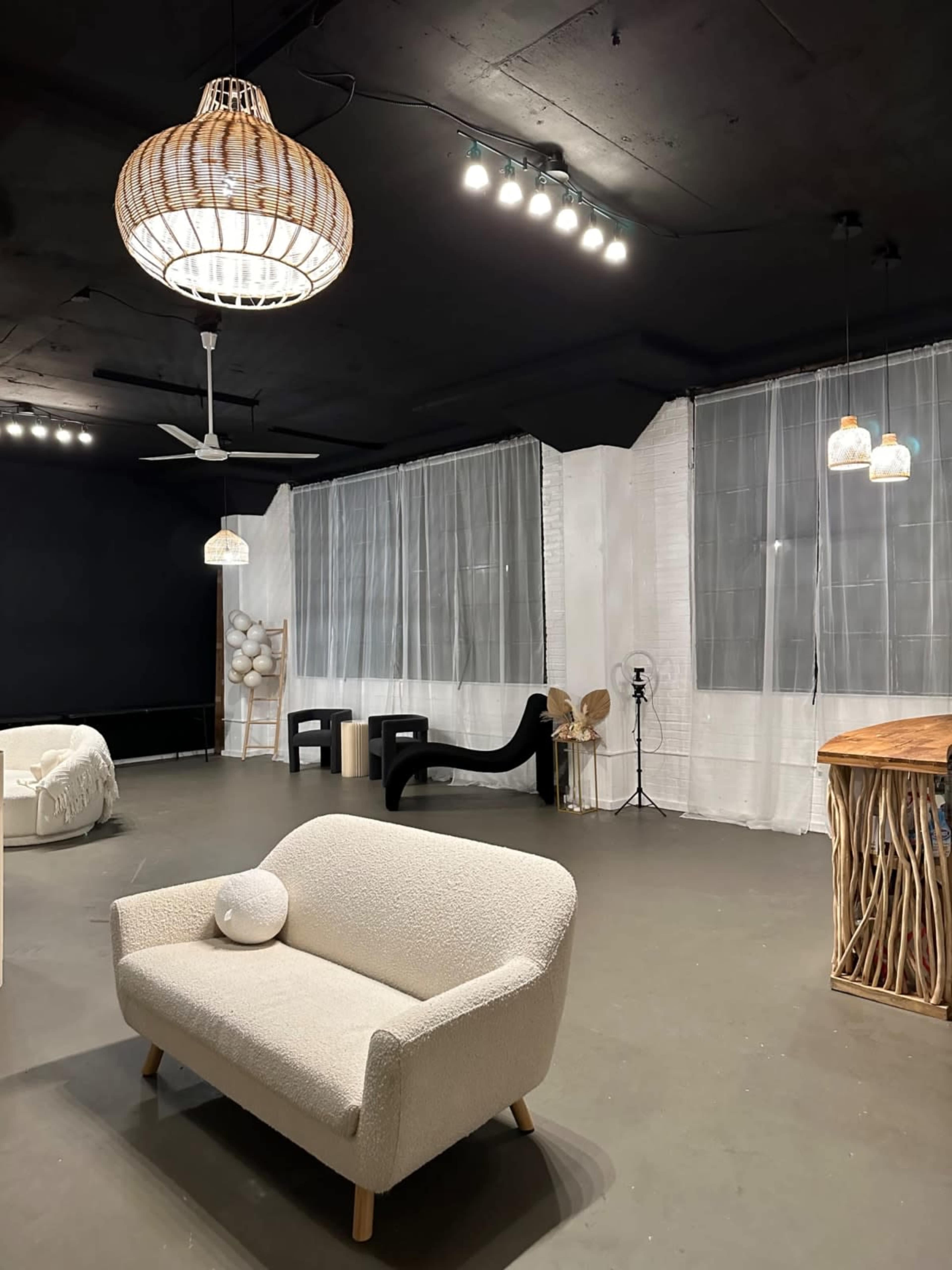 The image shows a spacious, minimalist room with a light-colored sofa, a large wooden table, and various pendant lights hanging from the ceiling.
