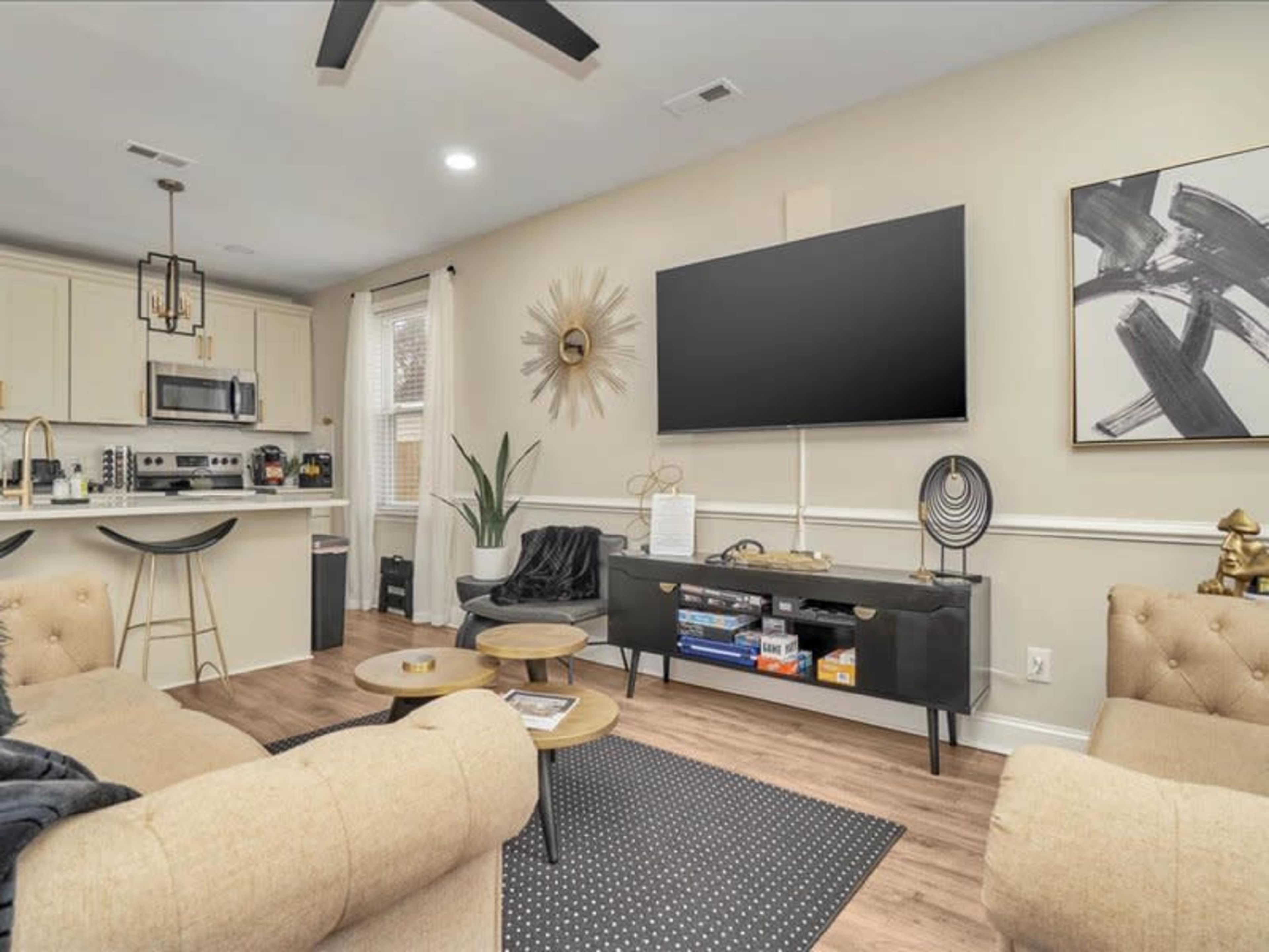 A modern living room with beige sofas, a dark rug, a large wall-mounted television, and a small dining area in the background.