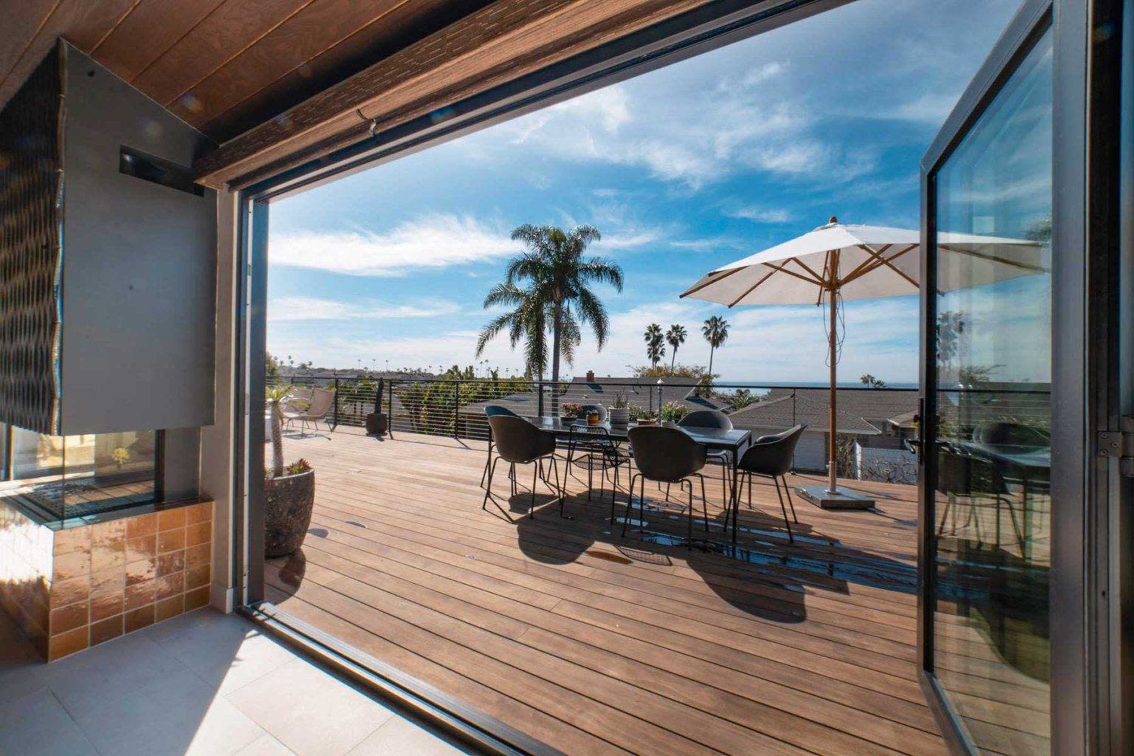 A spacious wooden deck with a dining table and chairs, surrounded by palm trees and an umbrella, under a bright blue sky.