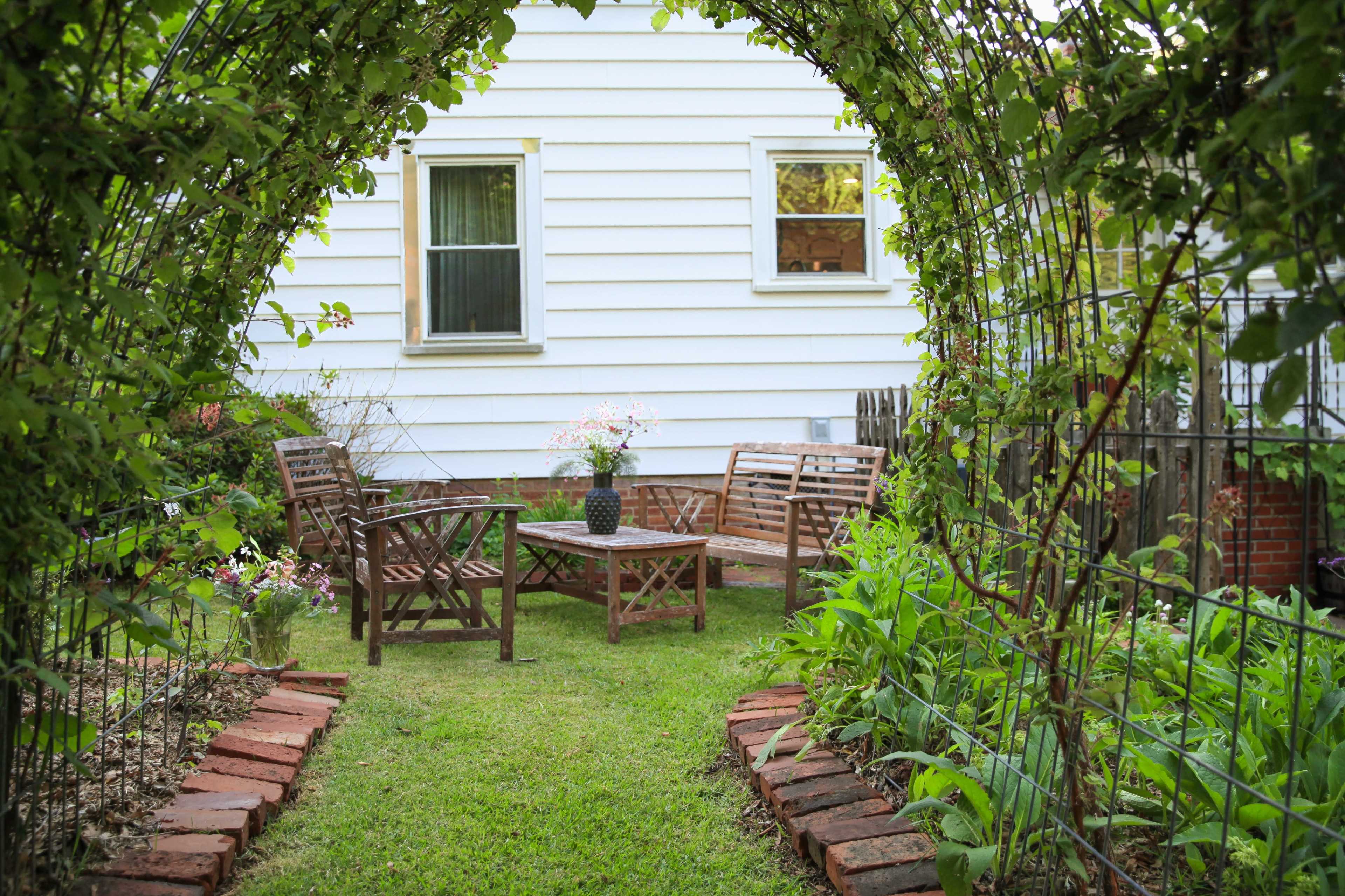 A small, green garden features a seating area with wooden chairs and a table, framed by an archway of foliage leading to a white house.