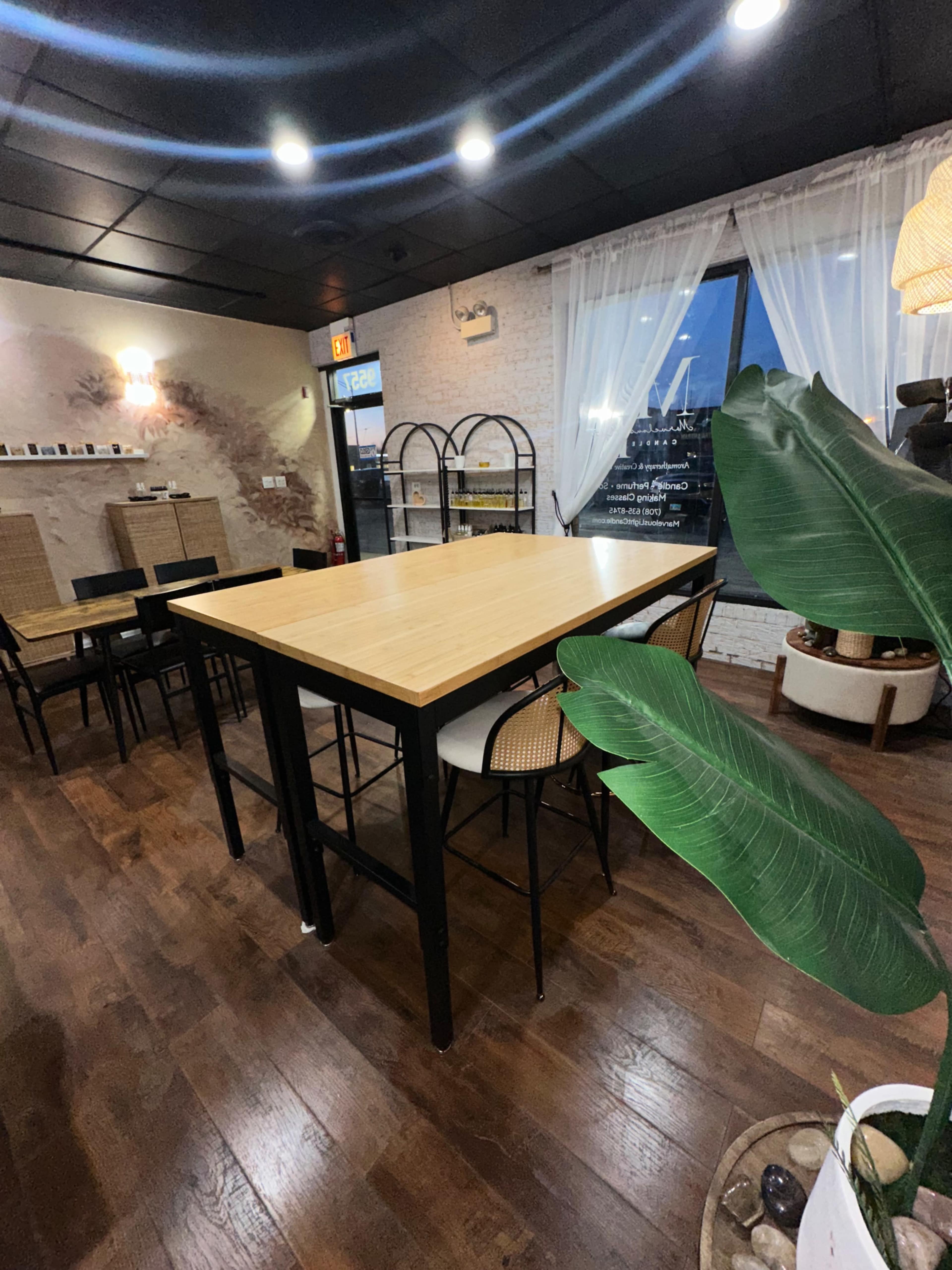 A spacious interior features a wooden table surrounded by matching high stools, with large windows allowing natural light to enter.