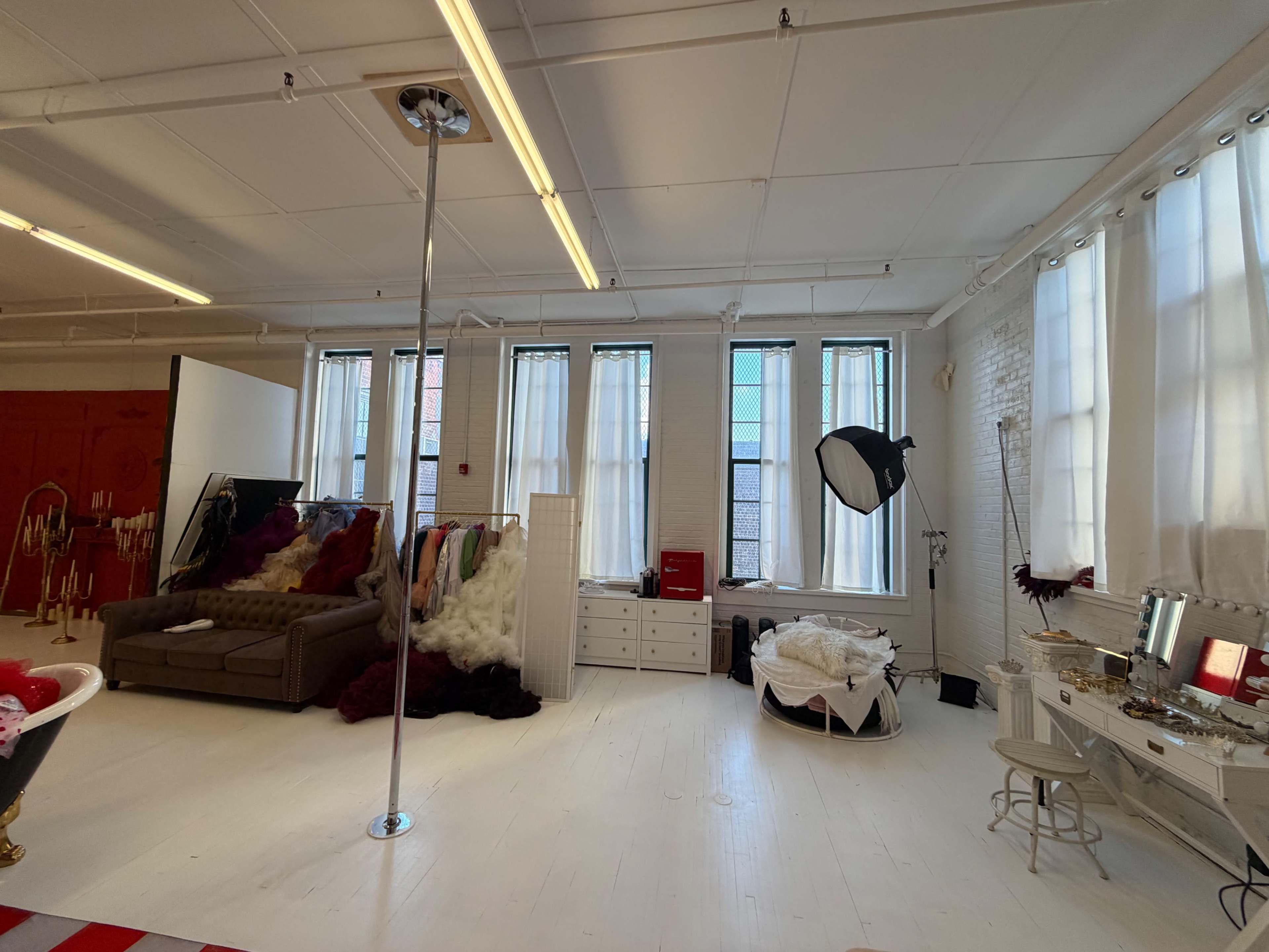The image shows a spacious, well-lit studio with large windows, a pole in the center, and various props and furniture, including a couch and mirrors.