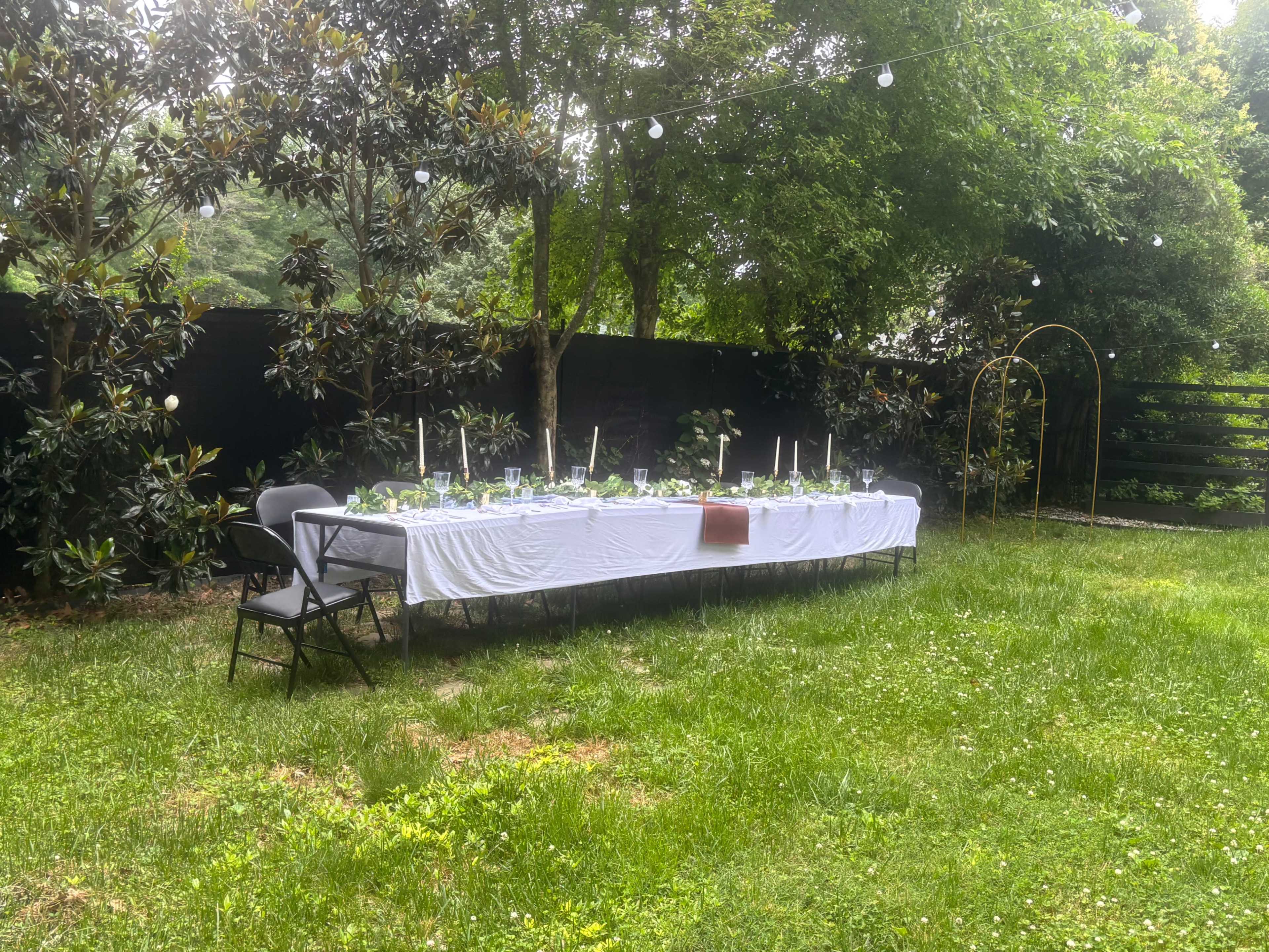 A long table adorned with white linens and candles is set up in a grassy area surrounded by trees and a dark fence.