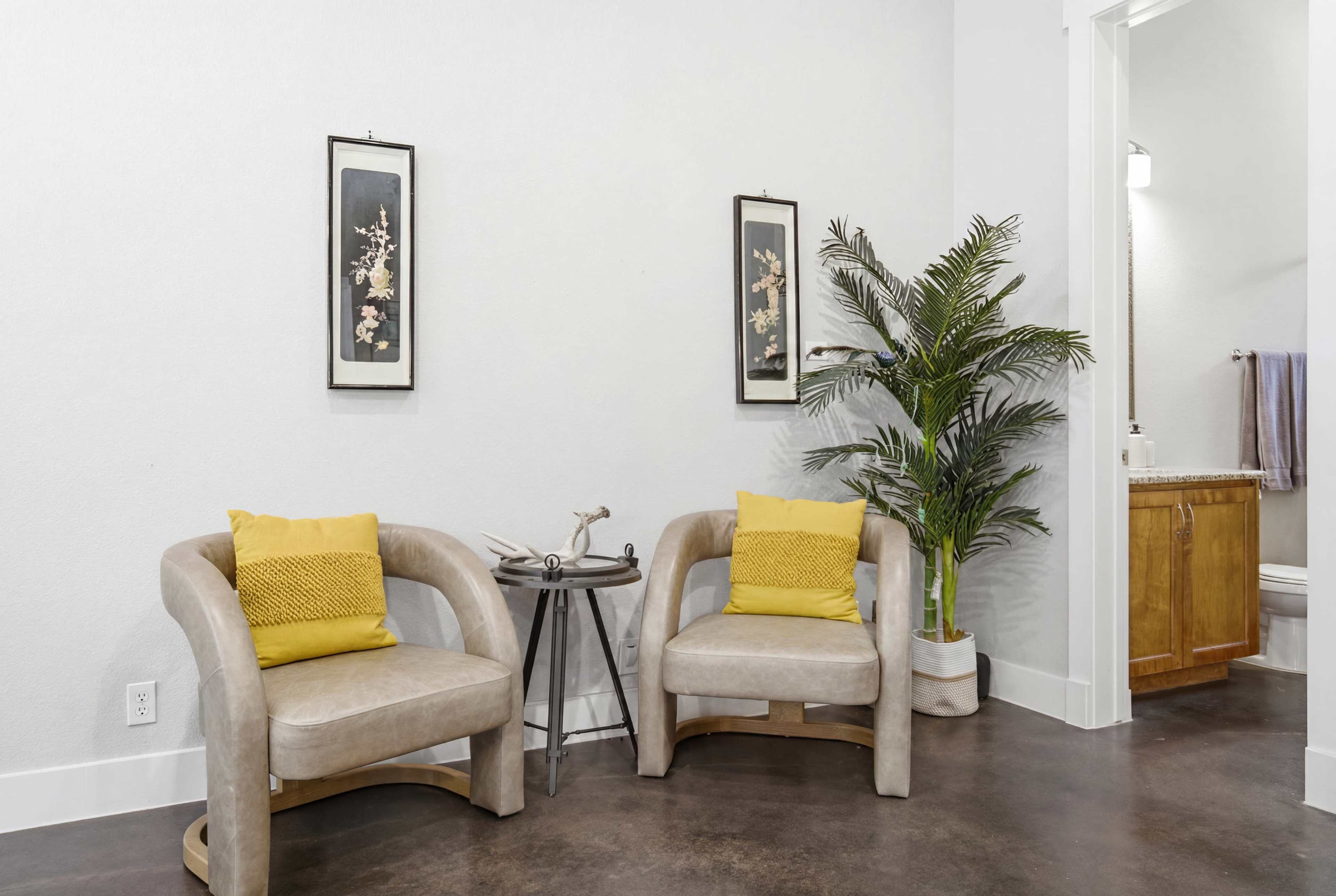The image shows a small seating area with two beige chairs, yellow cushions, a decorative plant, and framed artwork on the white walls.