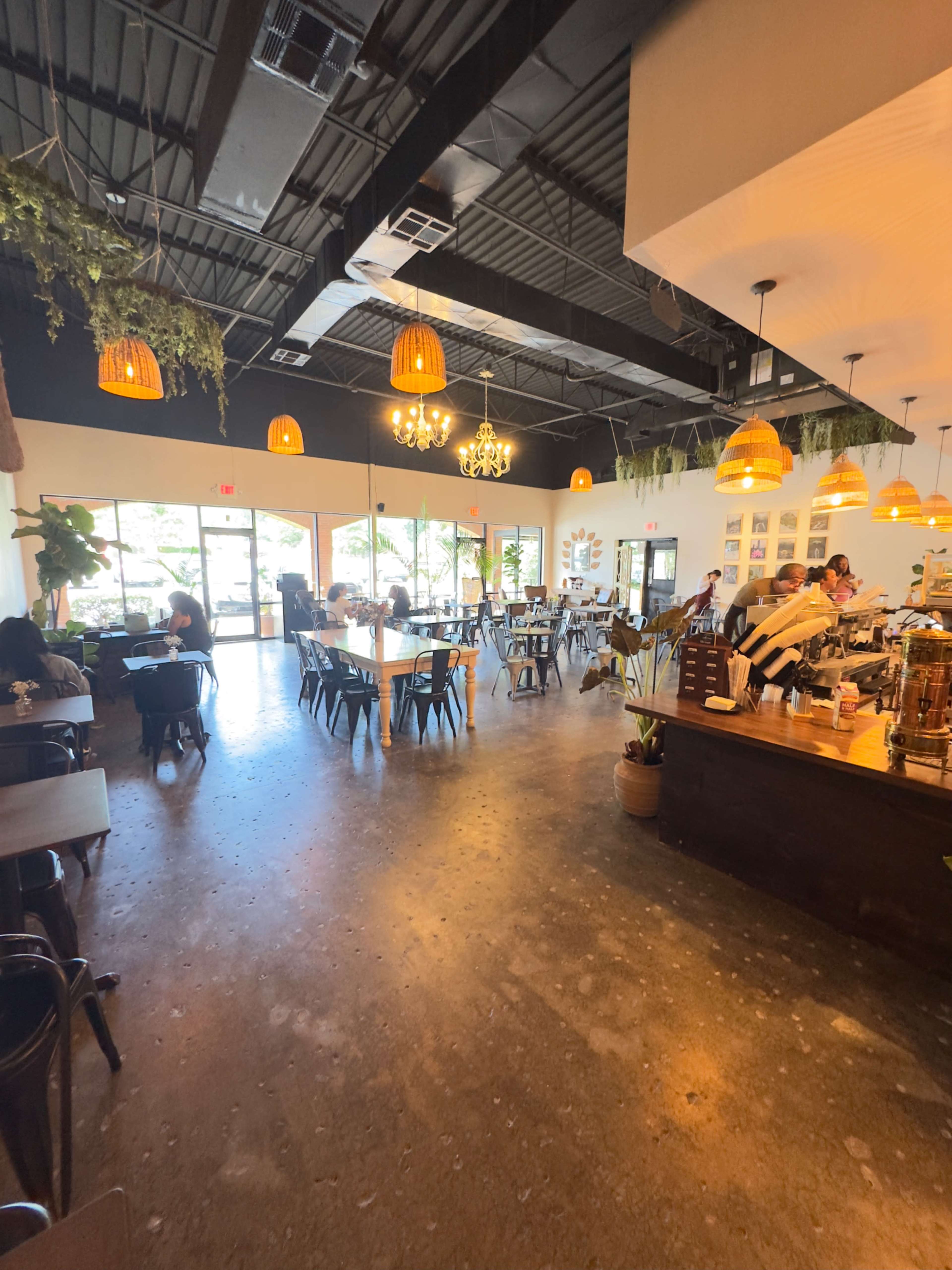 The image shows a spacious café interior with wooden furniture, pendant lighting, and large windows letting in natural light.