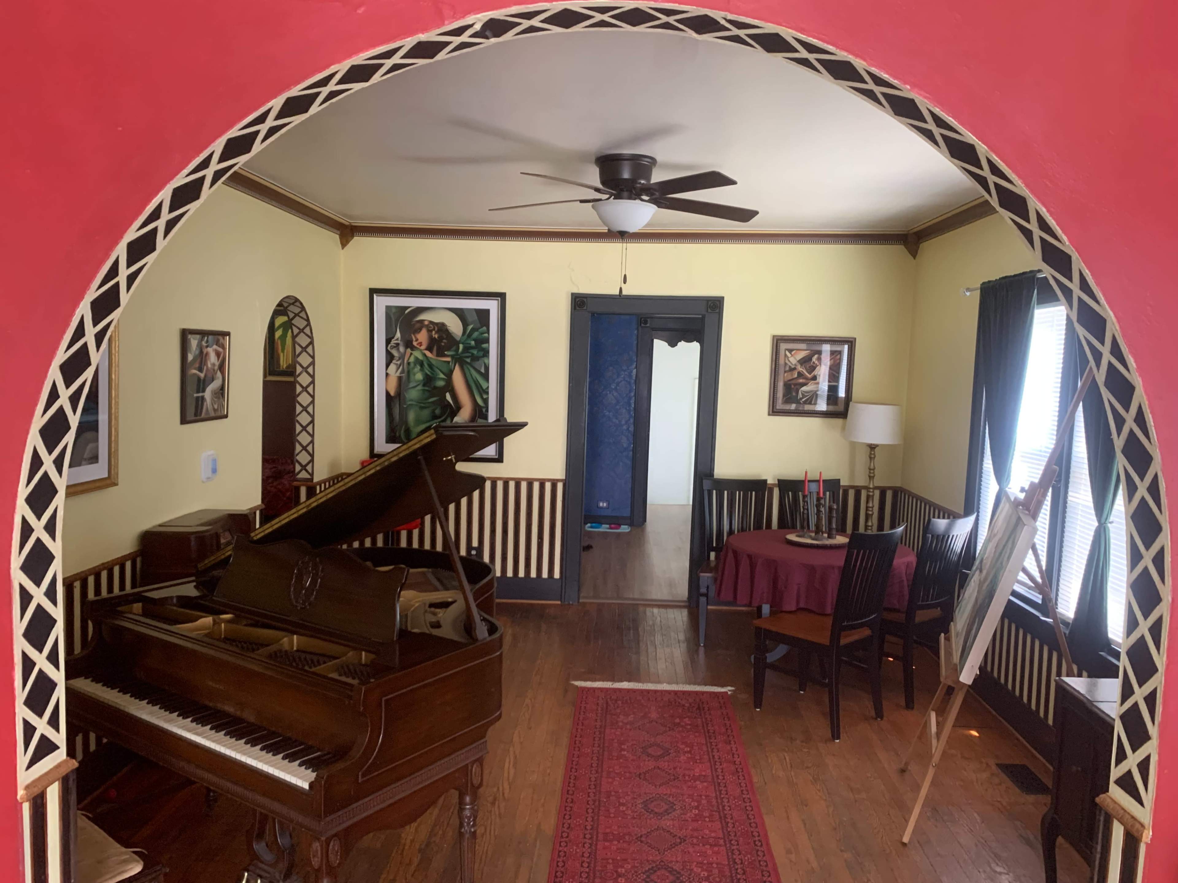 The image shows a room with a grand piano, a round table with chairs, and artwork on the walls, framed by a red archway.