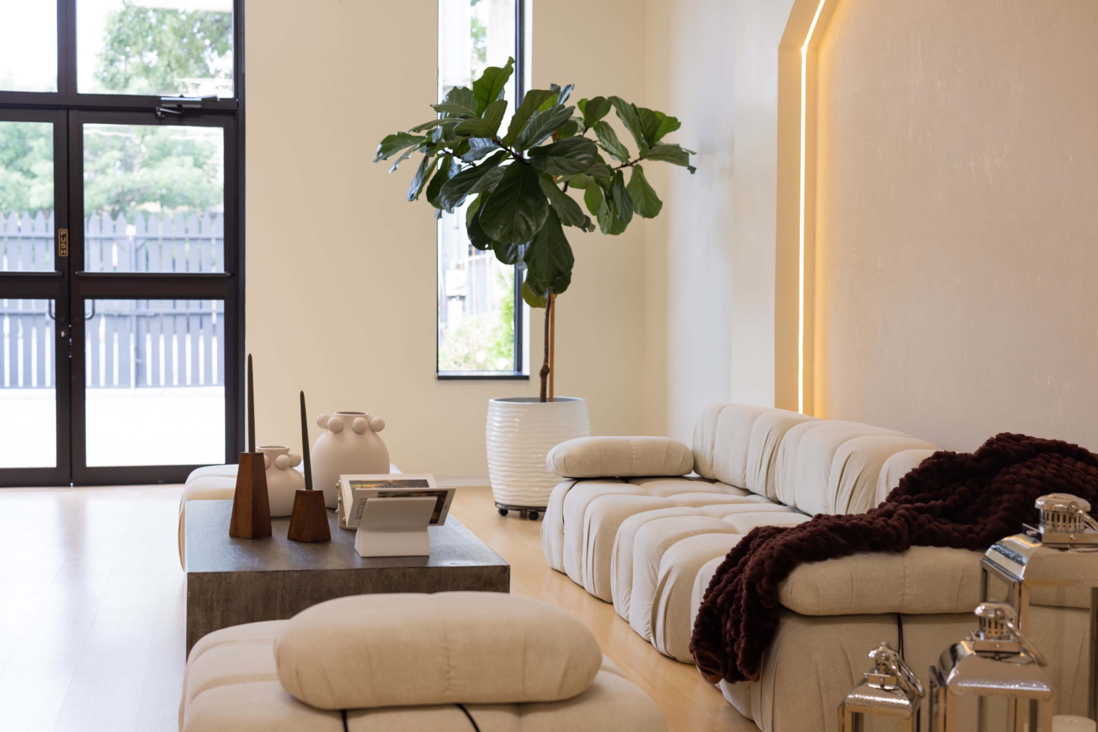 The image shows a minimalist living room with a light-colored sectional sofa, a coffee table, a decorative plant, and large windows letting in natural light.