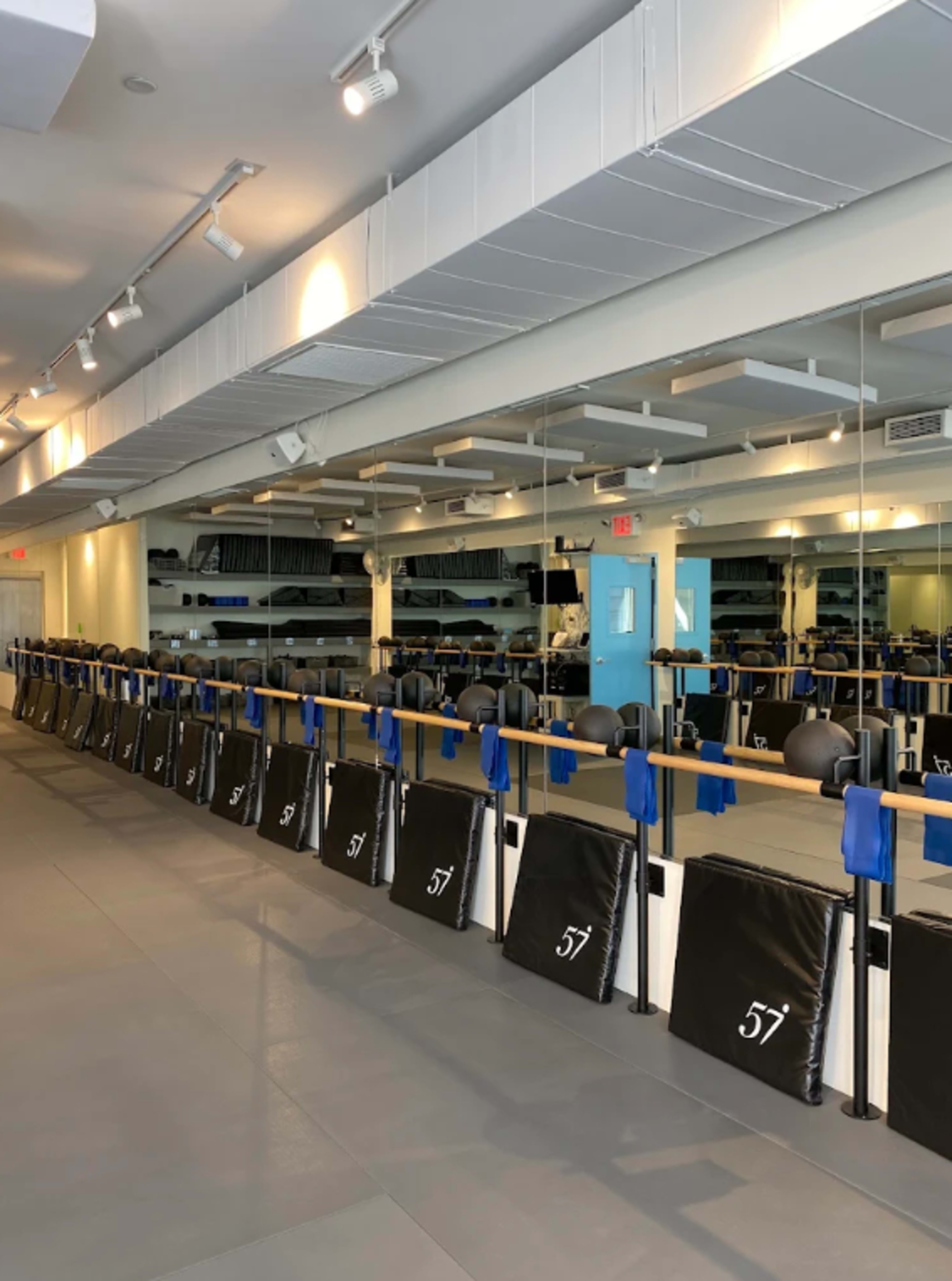 The image shows an exercise studio with a row of black barre pads and blue resistance bands set against a mirrored wall.