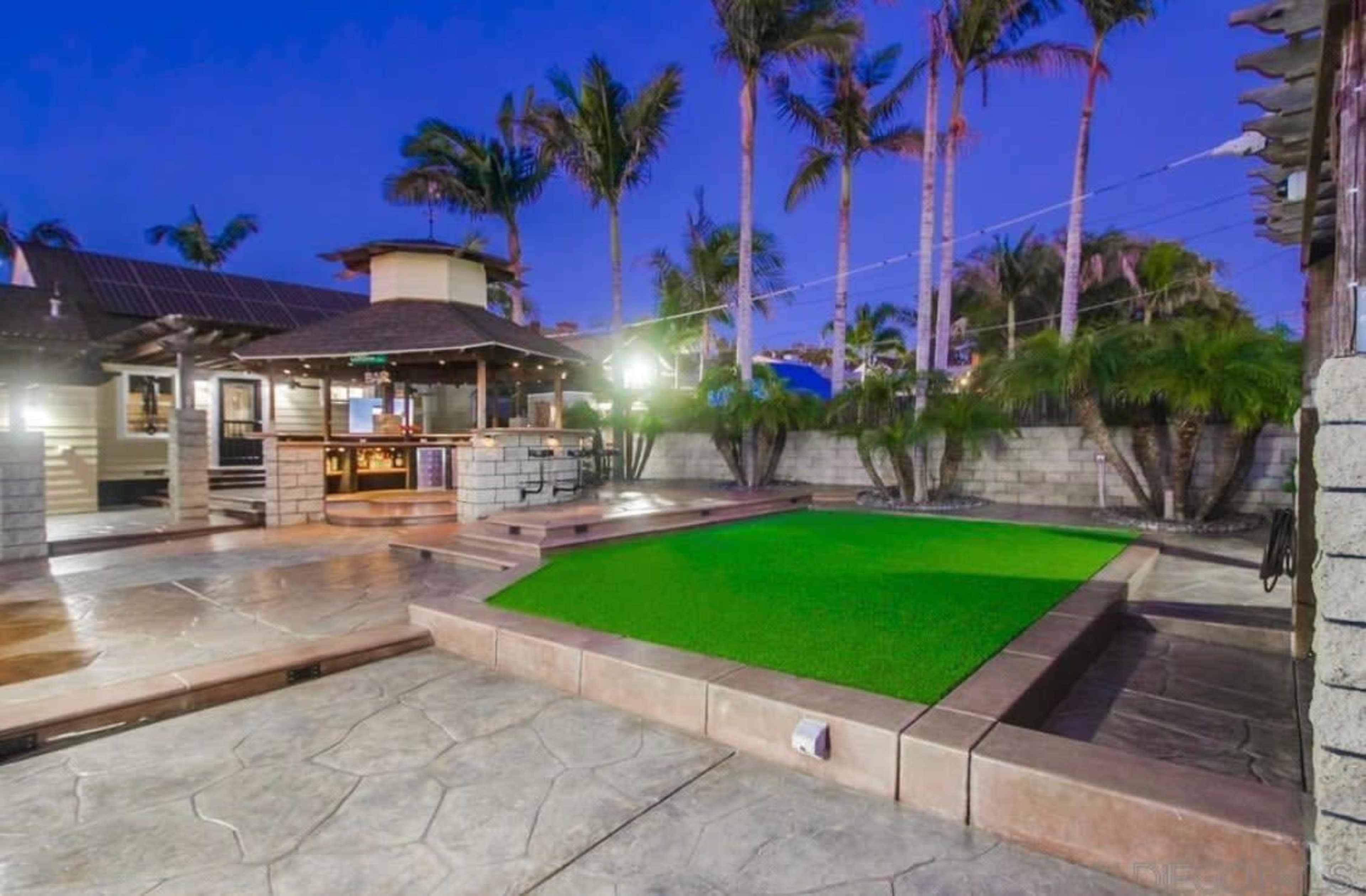 The image shows a landscaped outdoor area featuring a green synthetic lawn, stone pathways, palm trees, and a gazebo-like structure.