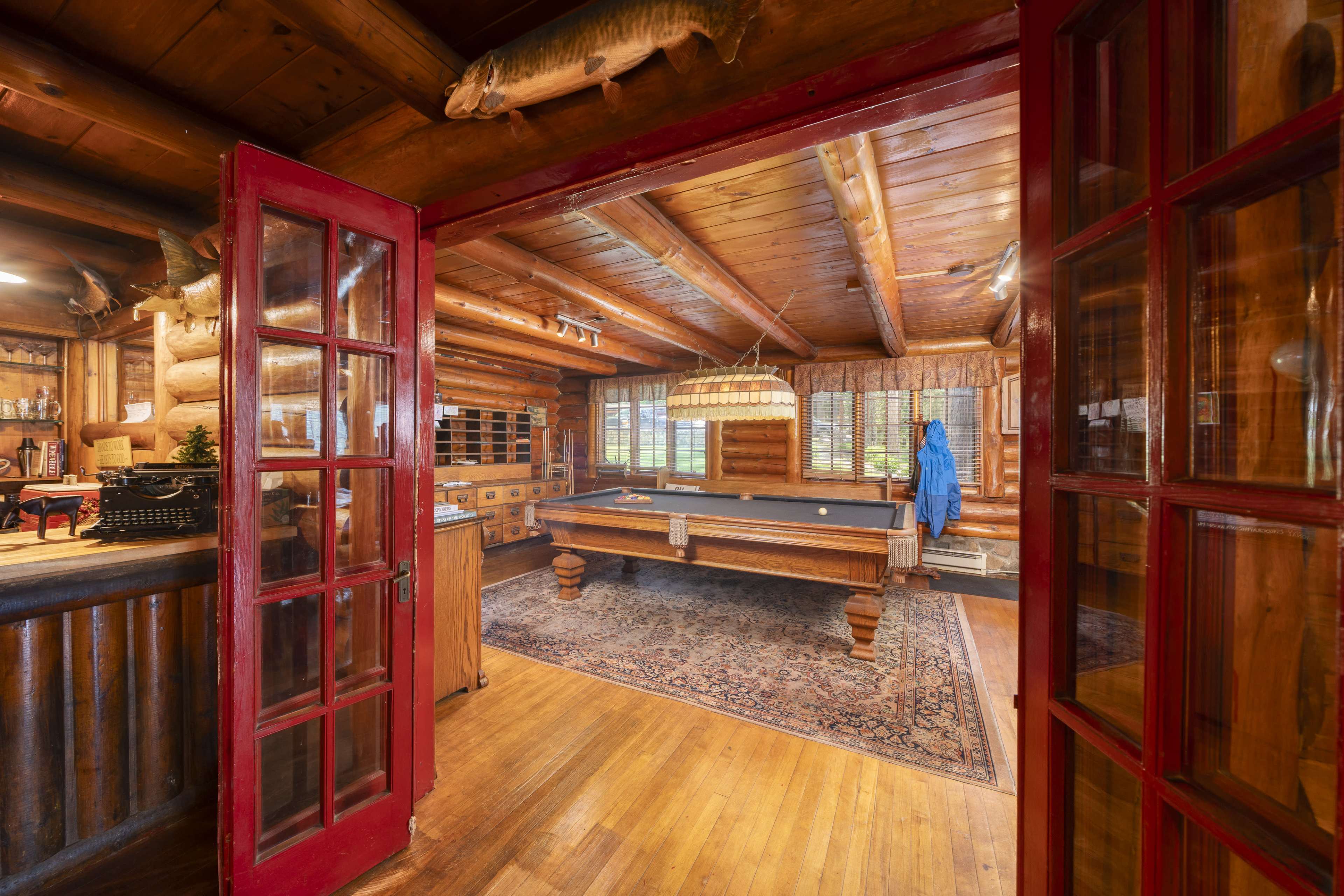 The image shows a rustic wooden interior of a cabin with a pool table, large windows, and a doorway leading to another room.
