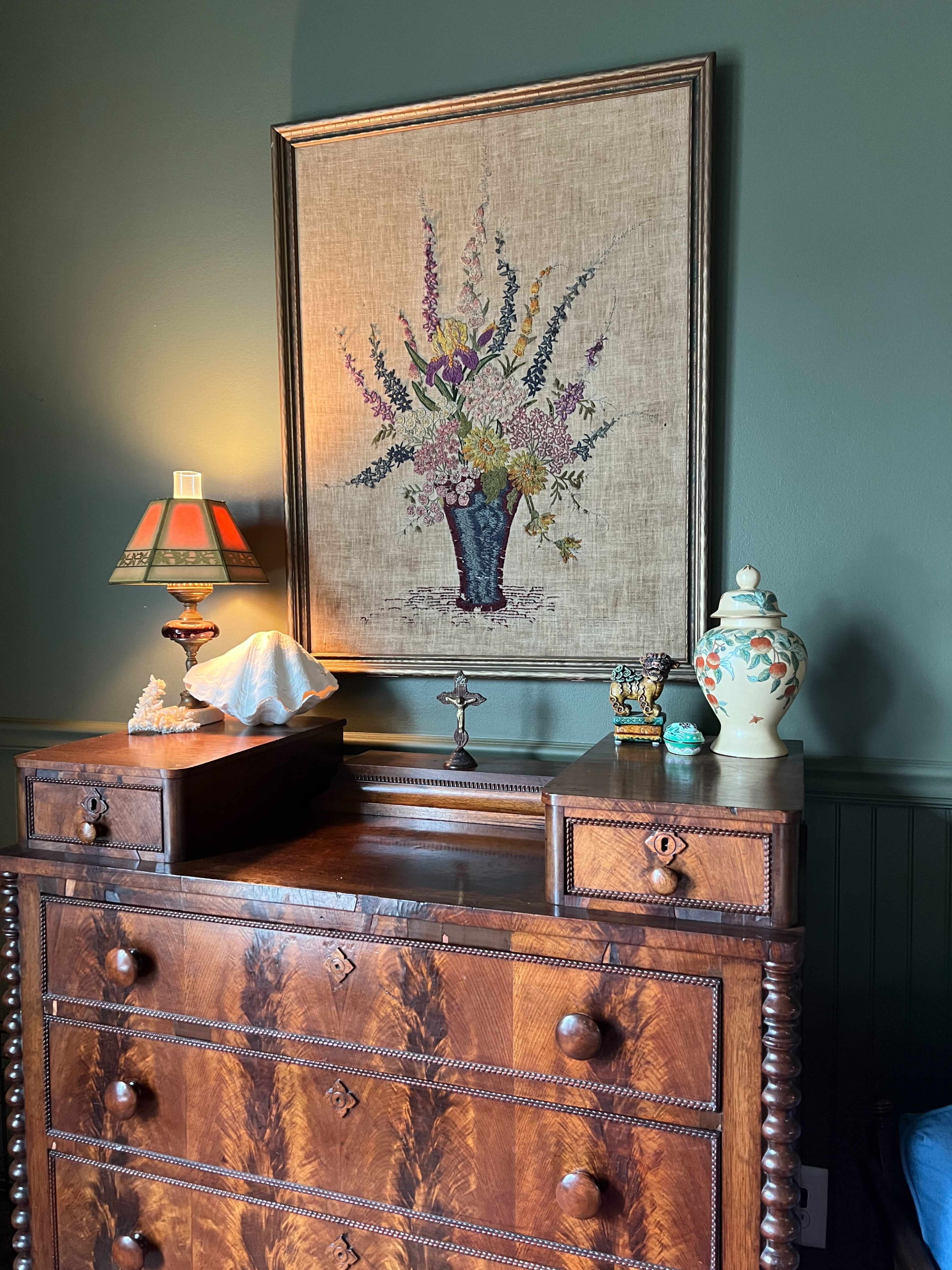 A wooden dresser with floral decor and a small lamp is positioned against a wall featuring a framed floral painting.