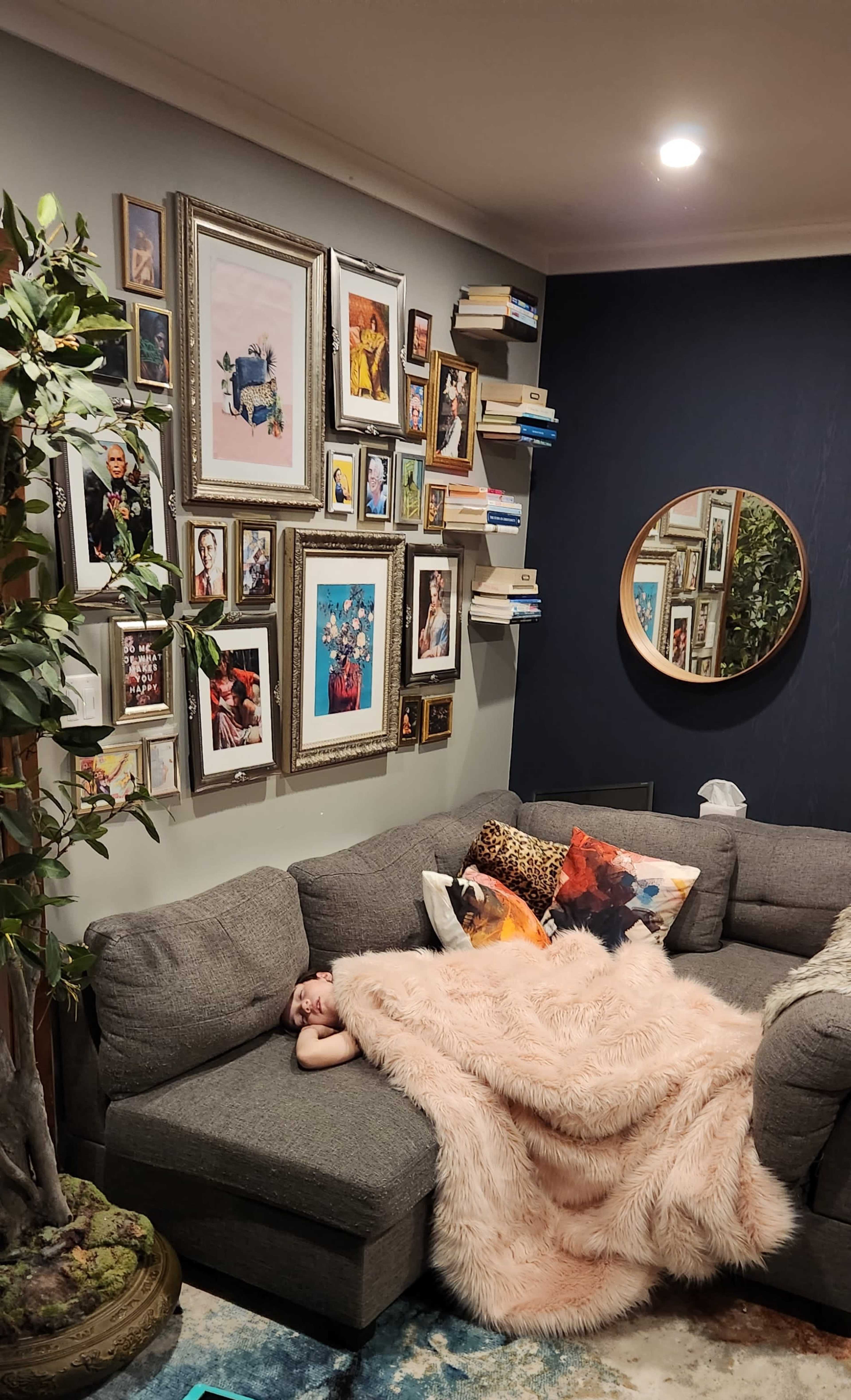 A person is sleeping on a gray sectional sofa draped with a pink blanket, surrounded by a gallery wall of framed photos and books.