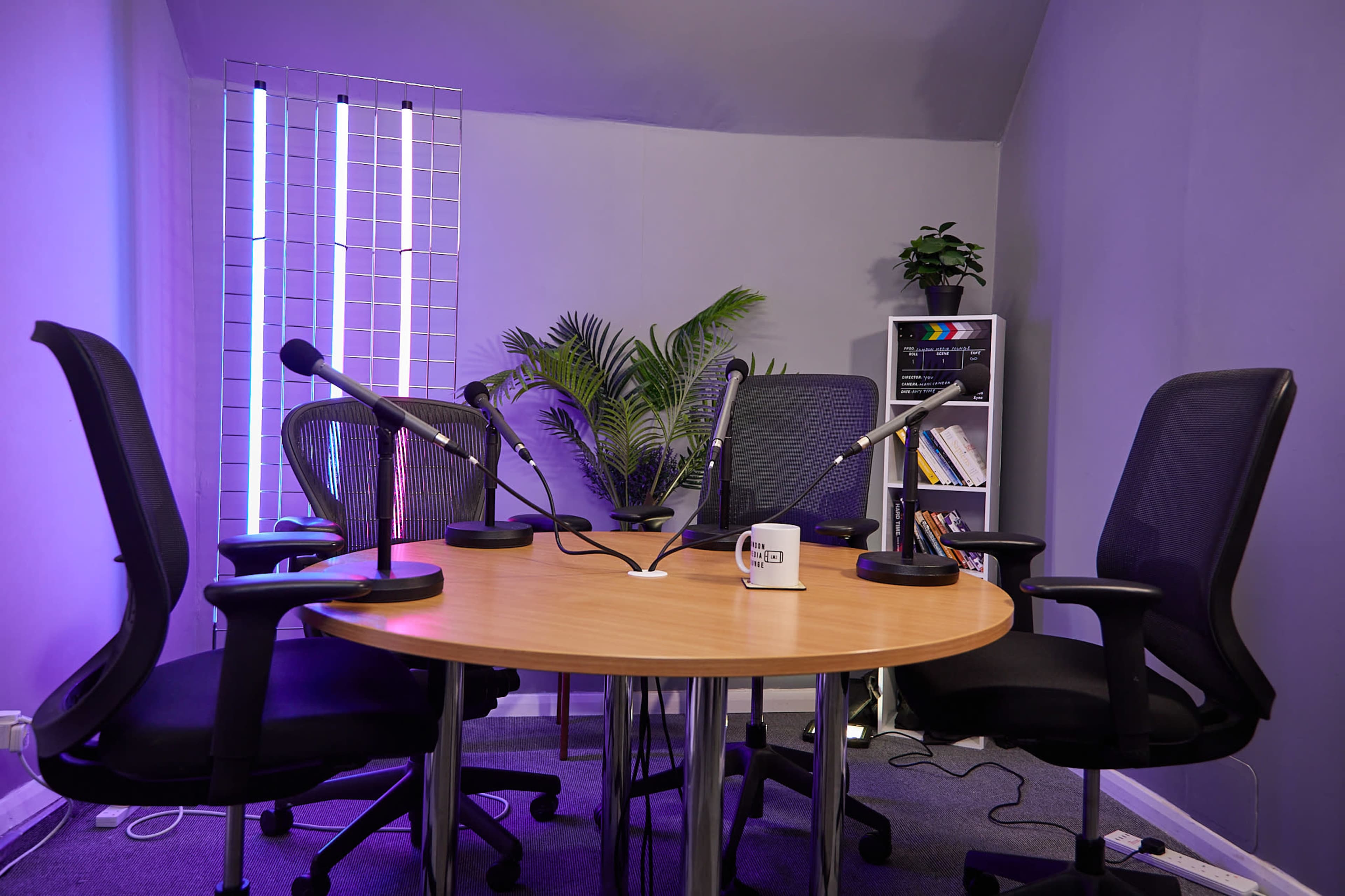 A round table with four black chairs is set in a gray room equipped with microphones and surrounded by plants and ambient lighting.