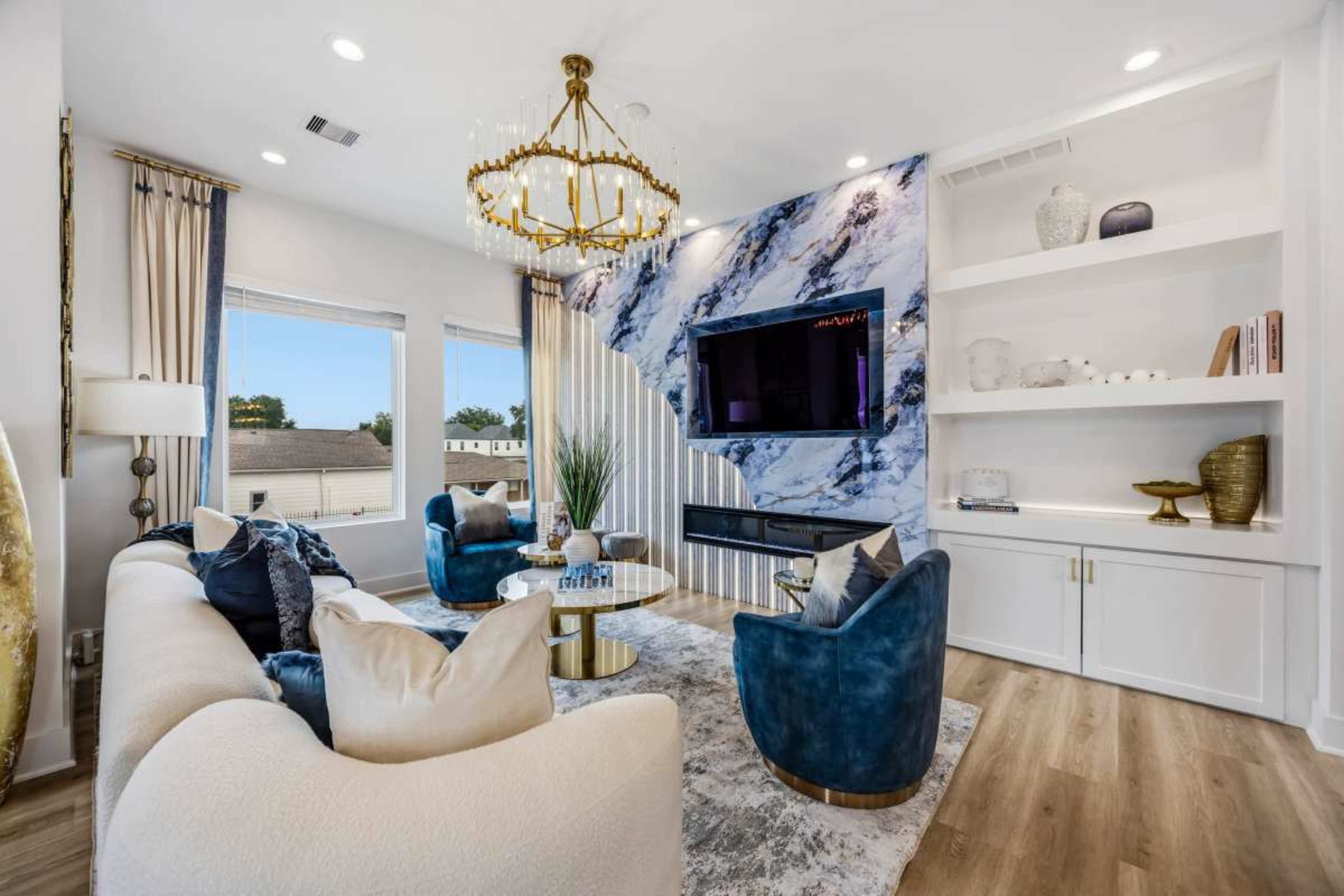 The image shows a modern living room featuring a curved sectional sofa, blue accent chairs, a round coffee table, and a large TV mounted on a wall with a marble design.