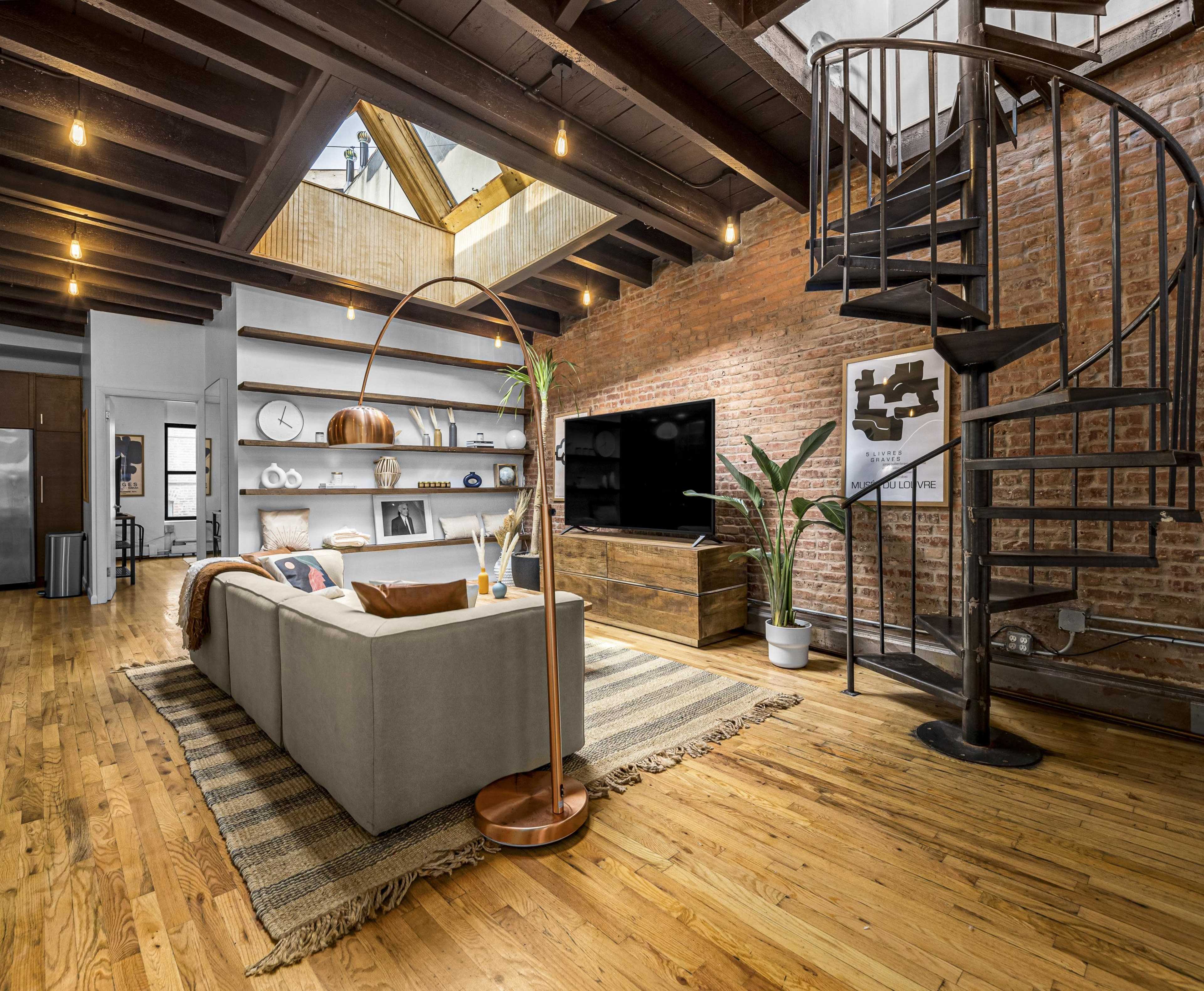 A spacious living room with exposed brick walls, a spiral staircase, wooden beams, and a sectional sofa arranged around a coffee table.