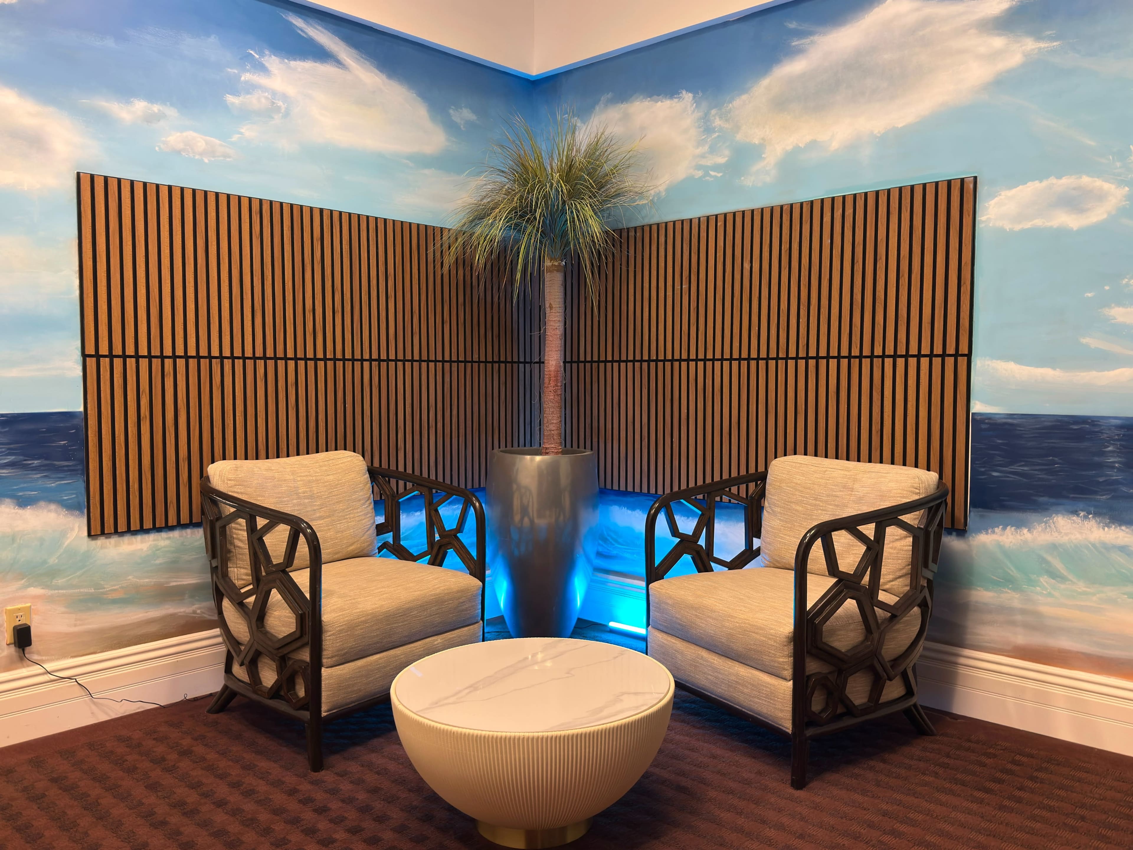 The image features a cozy seating area with two chairs facing each other, a round coffee table in the center, and a potted palm tree against a backdrop of a painted ocean scene.
