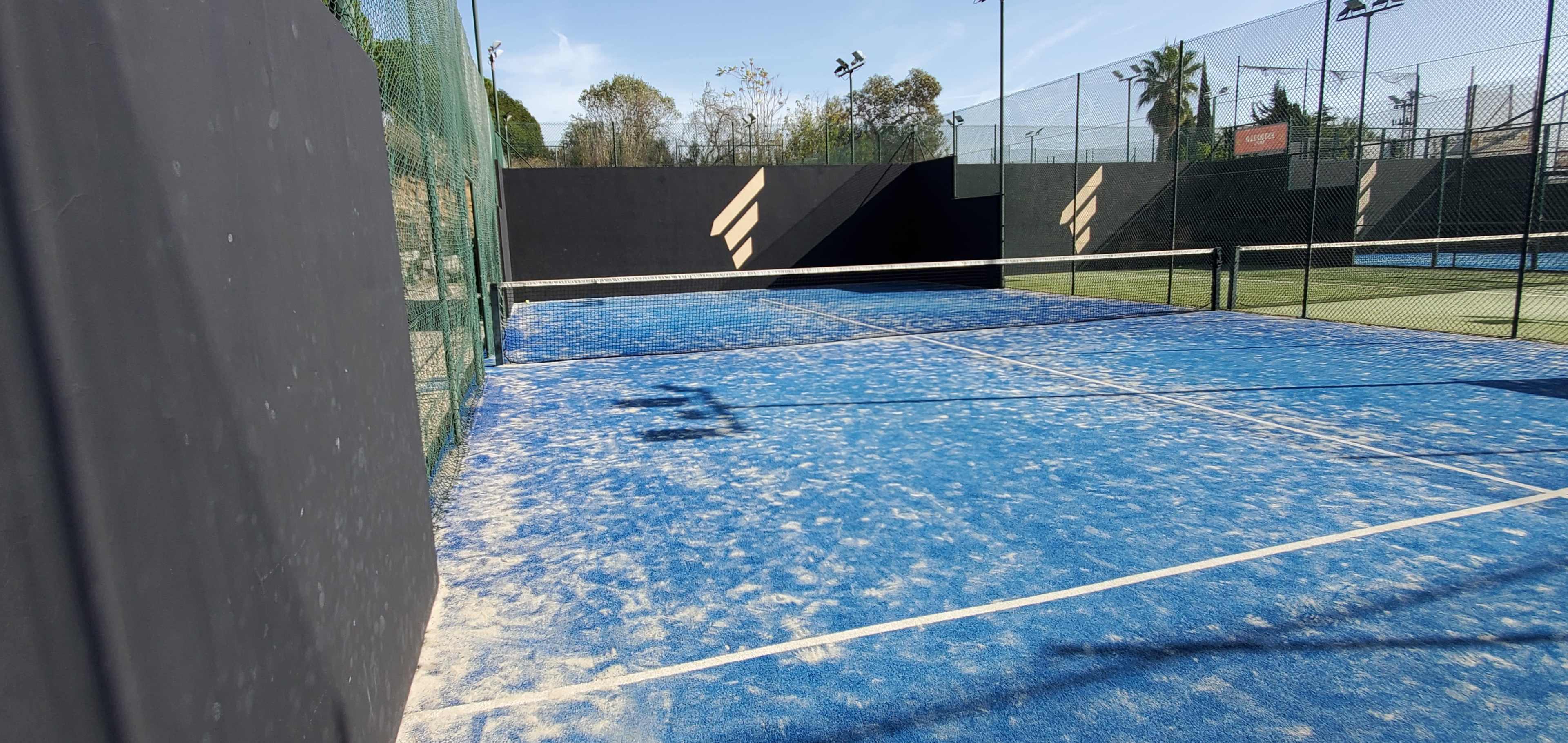 A blue tennis court is enclosed by a black wall and a green fence, with sunlight casting shadows on the surface.