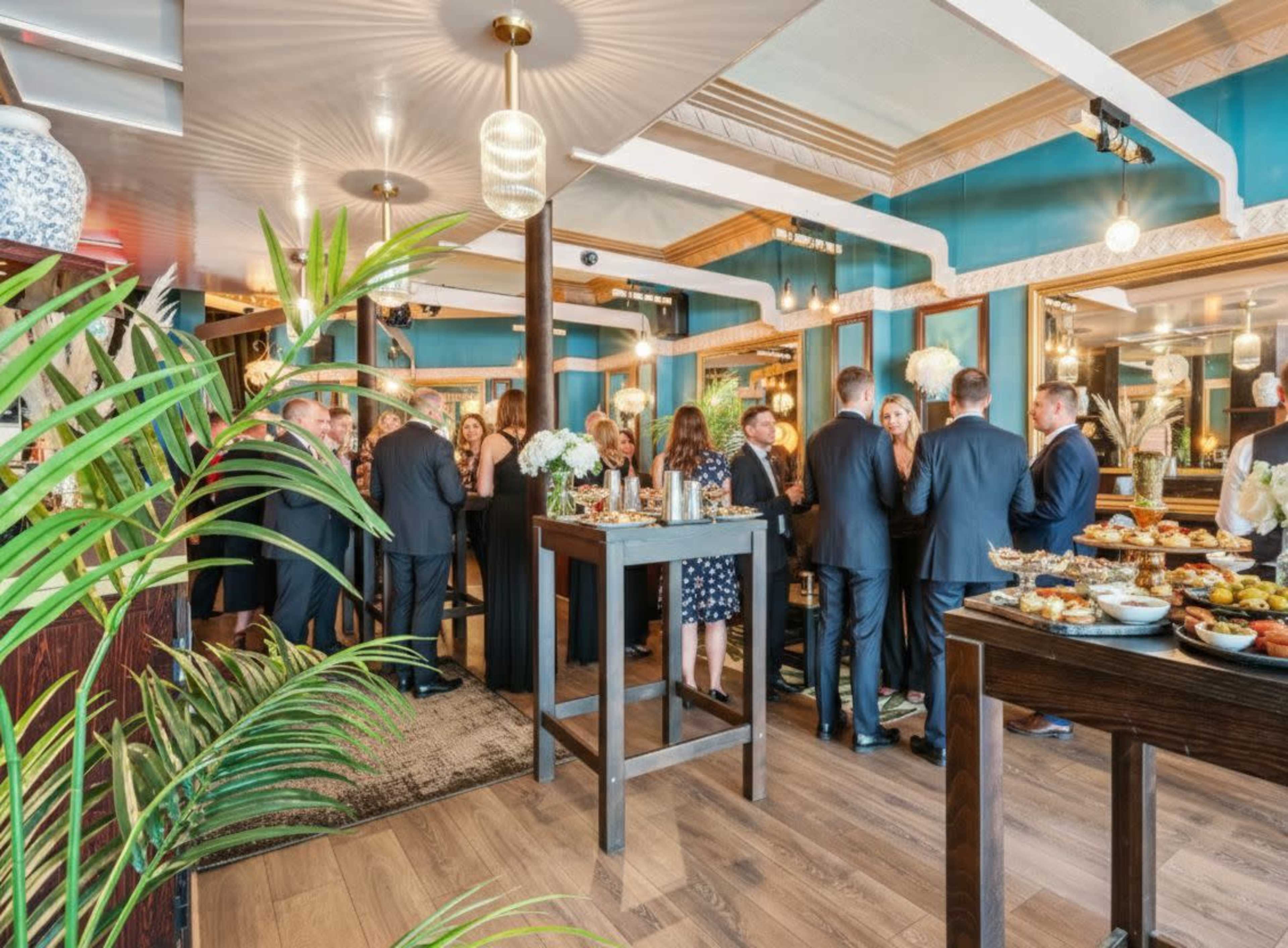 A group of people in formal attire is socializing at a stylish indoor gathering, surrounded by decorative plants and an assortment of food on tables.