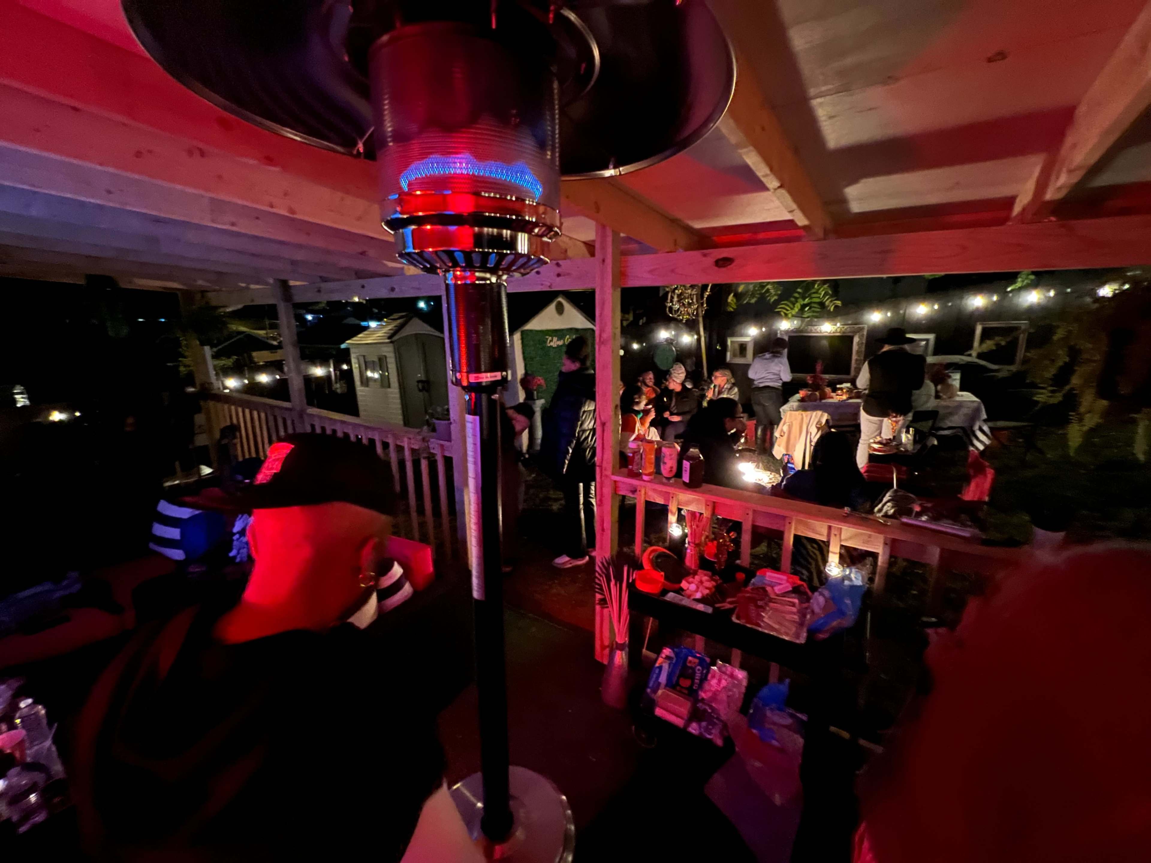A gathering on a wooden patio, with people seated around tables and a patio heater illuminating the scene under string lights.