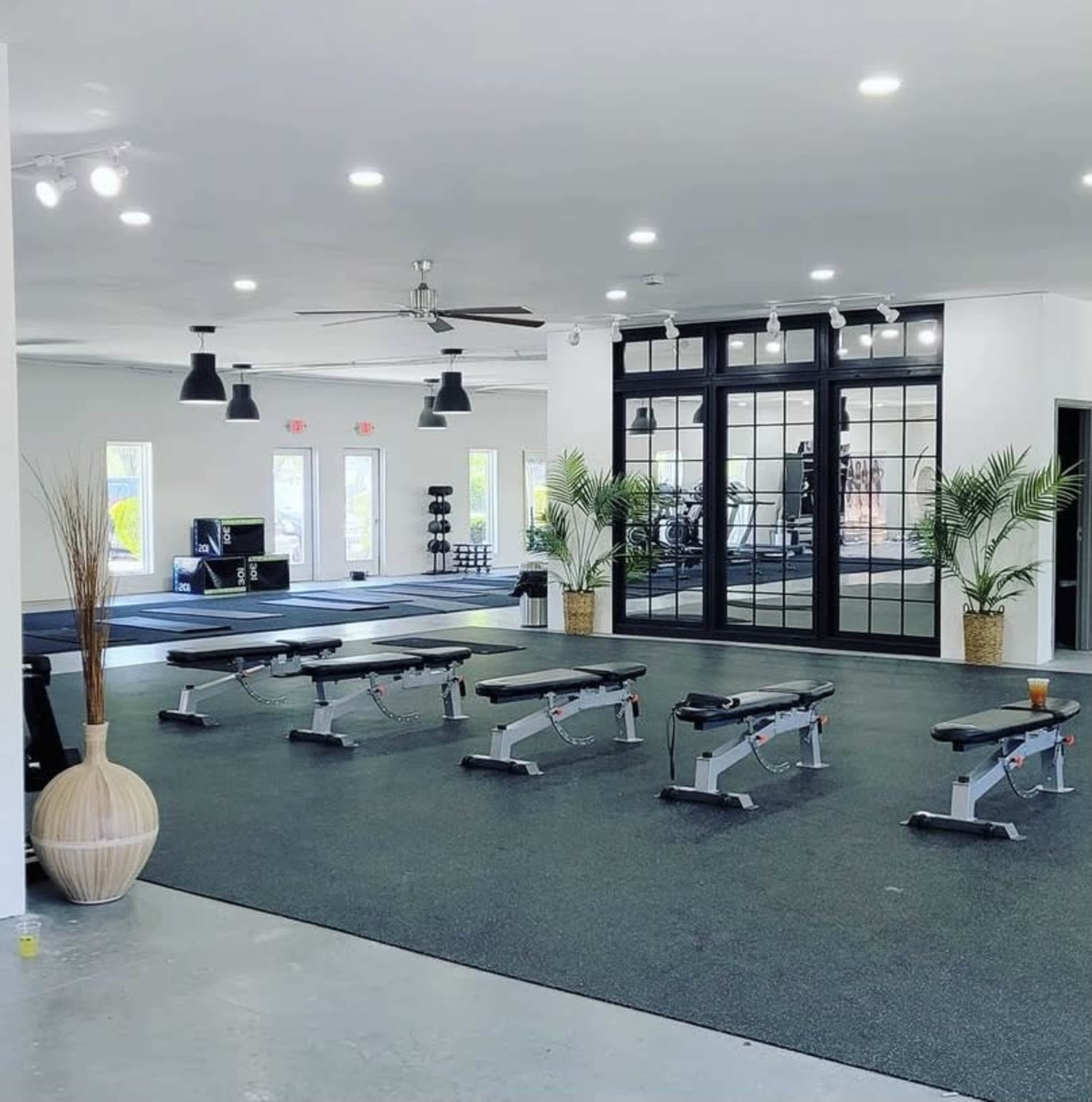 A modern gym with workout benches arranged on a rubber floor, large windows, and fitness equipment in the background.