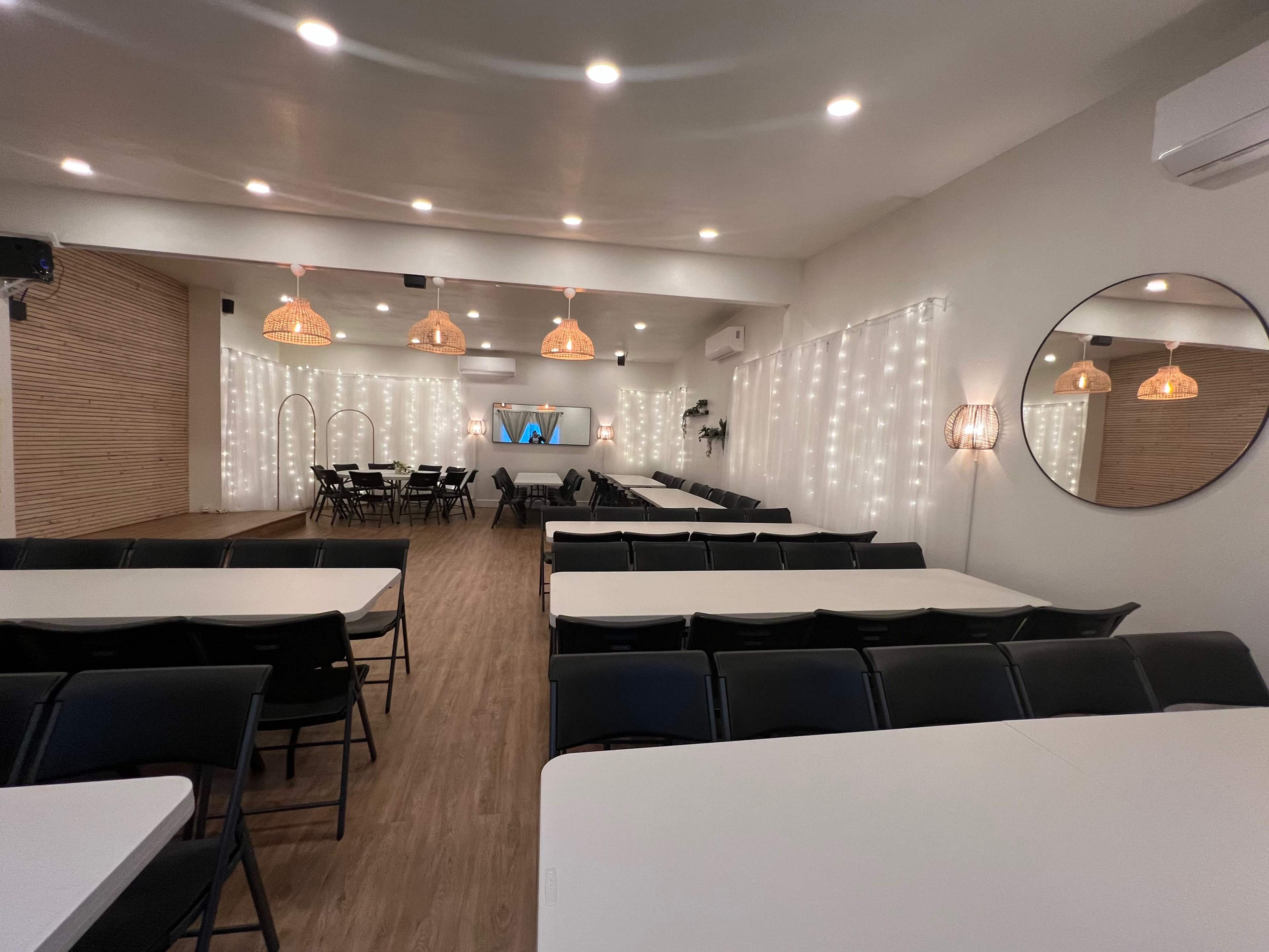 A spacious room is arranged with rows of tables and chairs, illuminated by pendant lights and adorned with string lights on the walls.