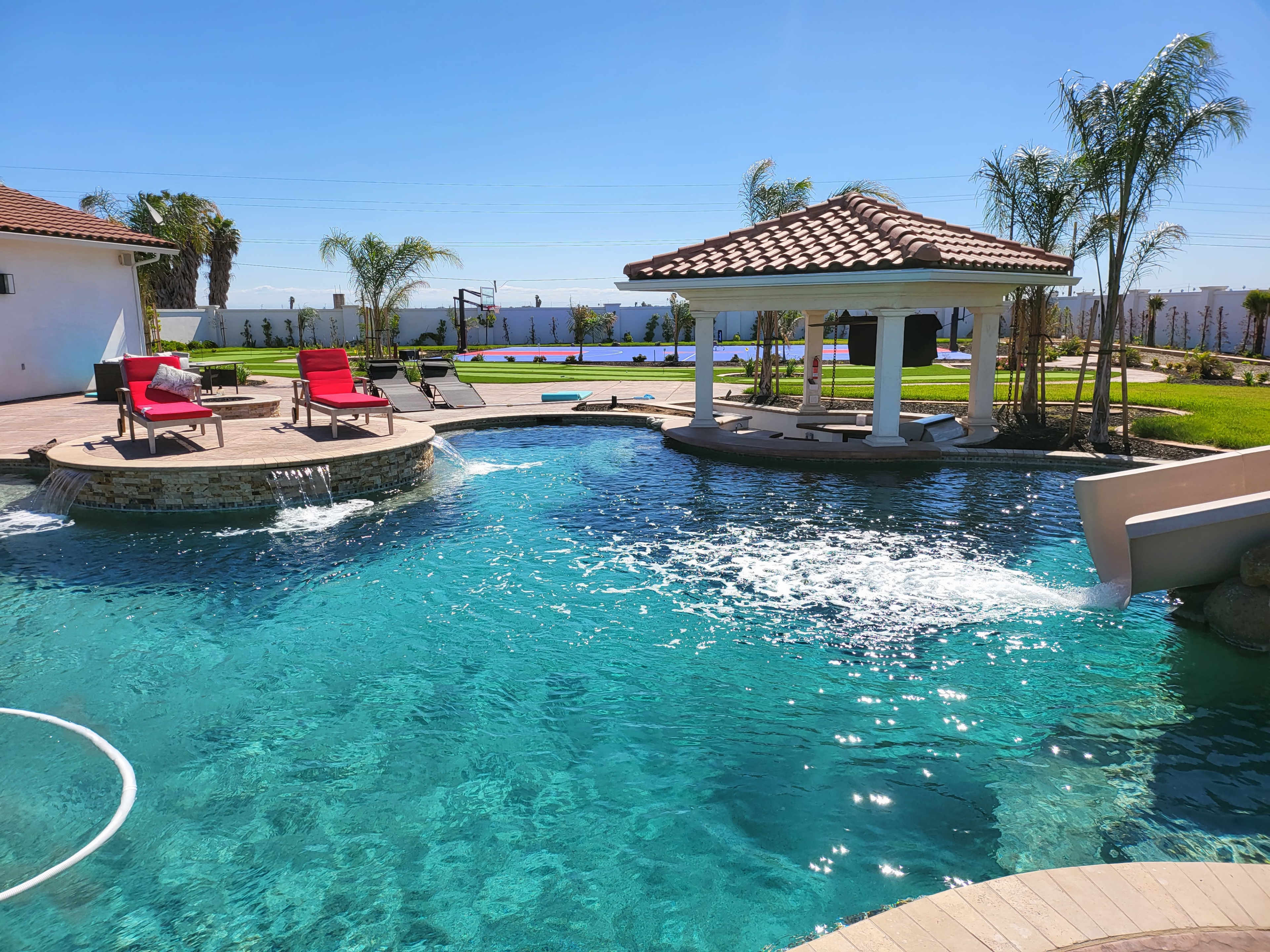 A backyard with a swimming pool featuring a fountain, lounge chairs, and a gazebo surrounded by palm trees and landscaping.