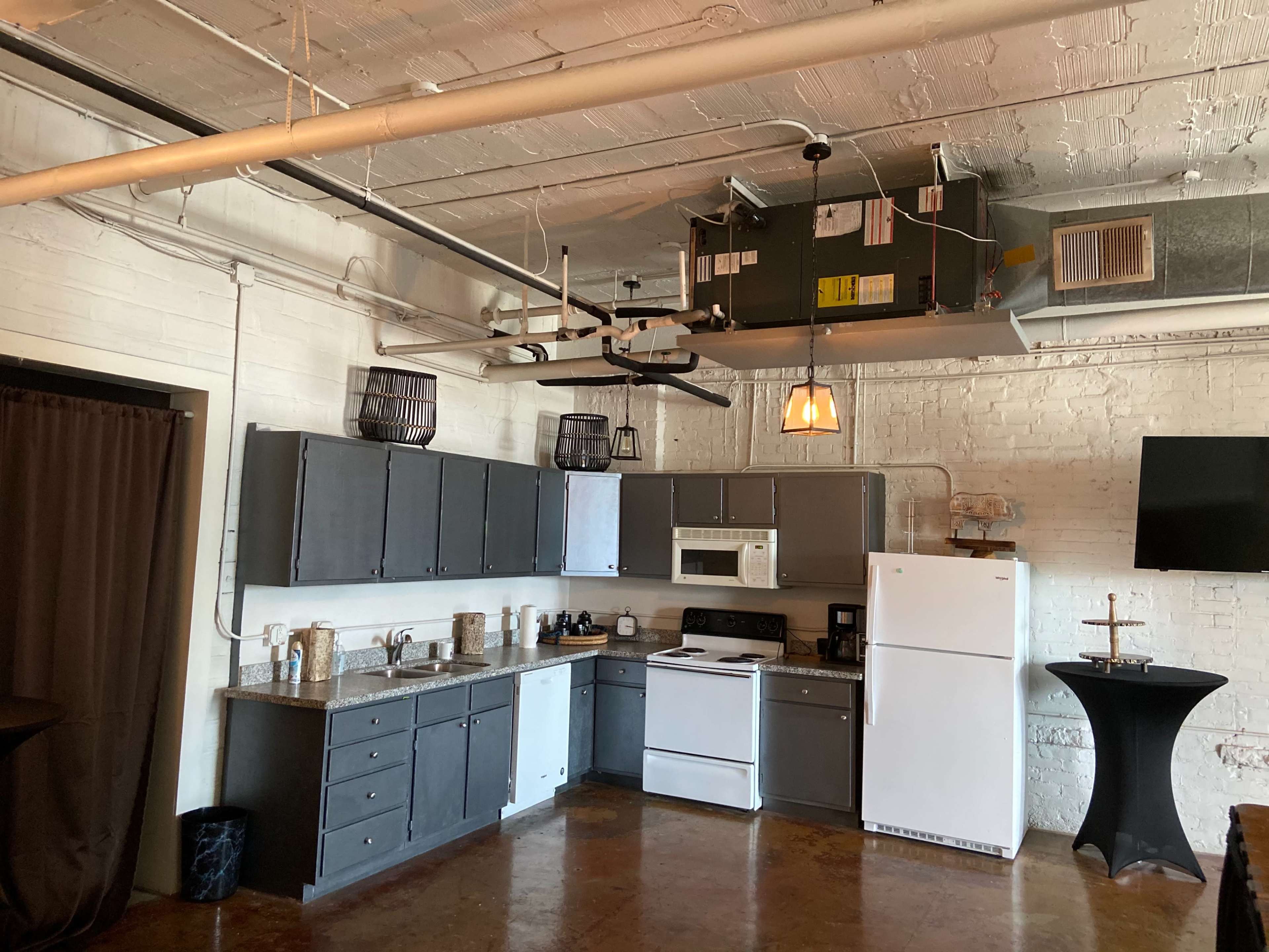 A kitchen with dark cabinetry, white appliances, and an island in a loft-style space with exposed brick walls and industrial elements.