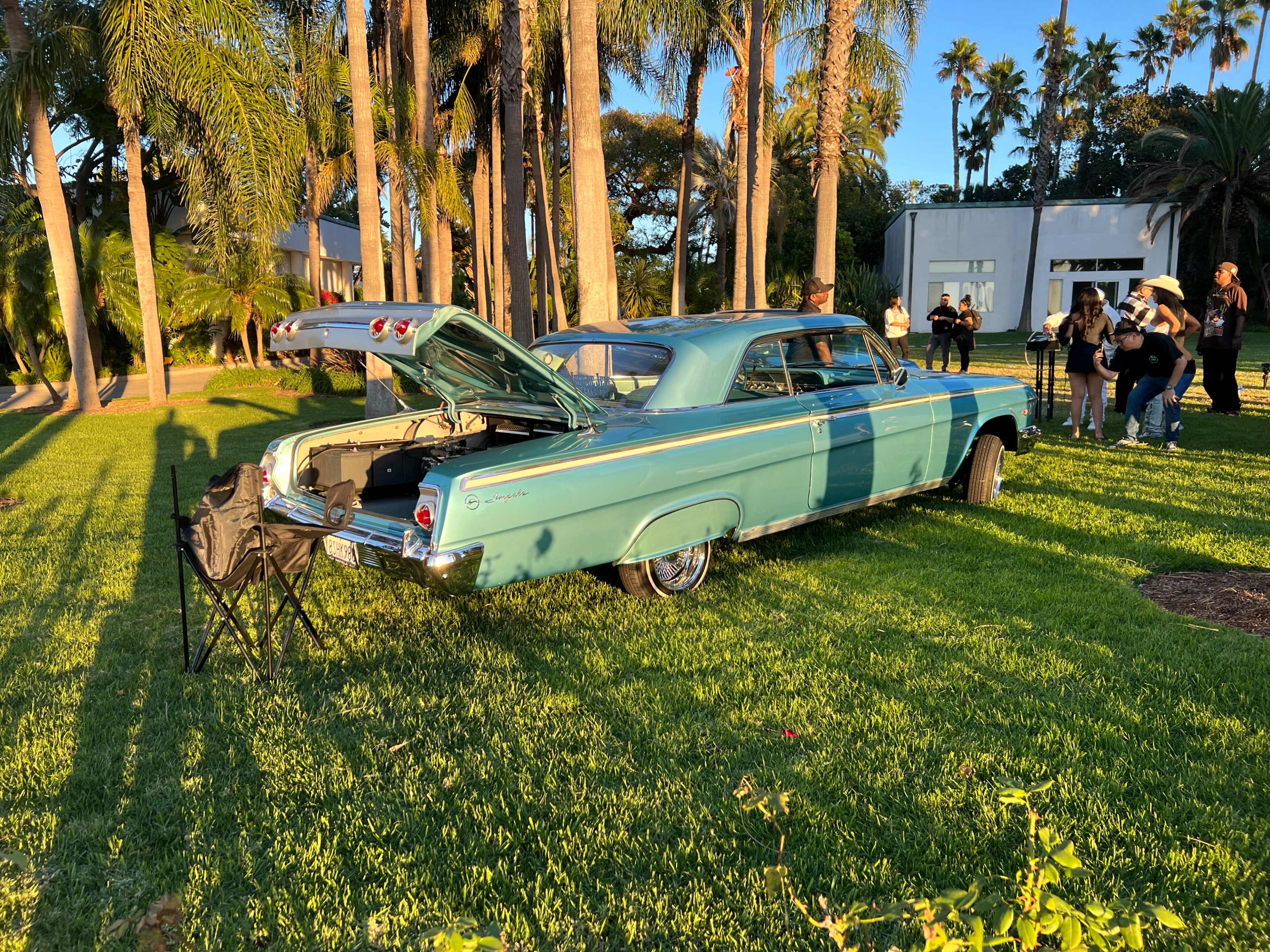 A vintage blue car with its trunk open is parked on a grassy area surrounded by palm trees, while a group of people stands nearby.