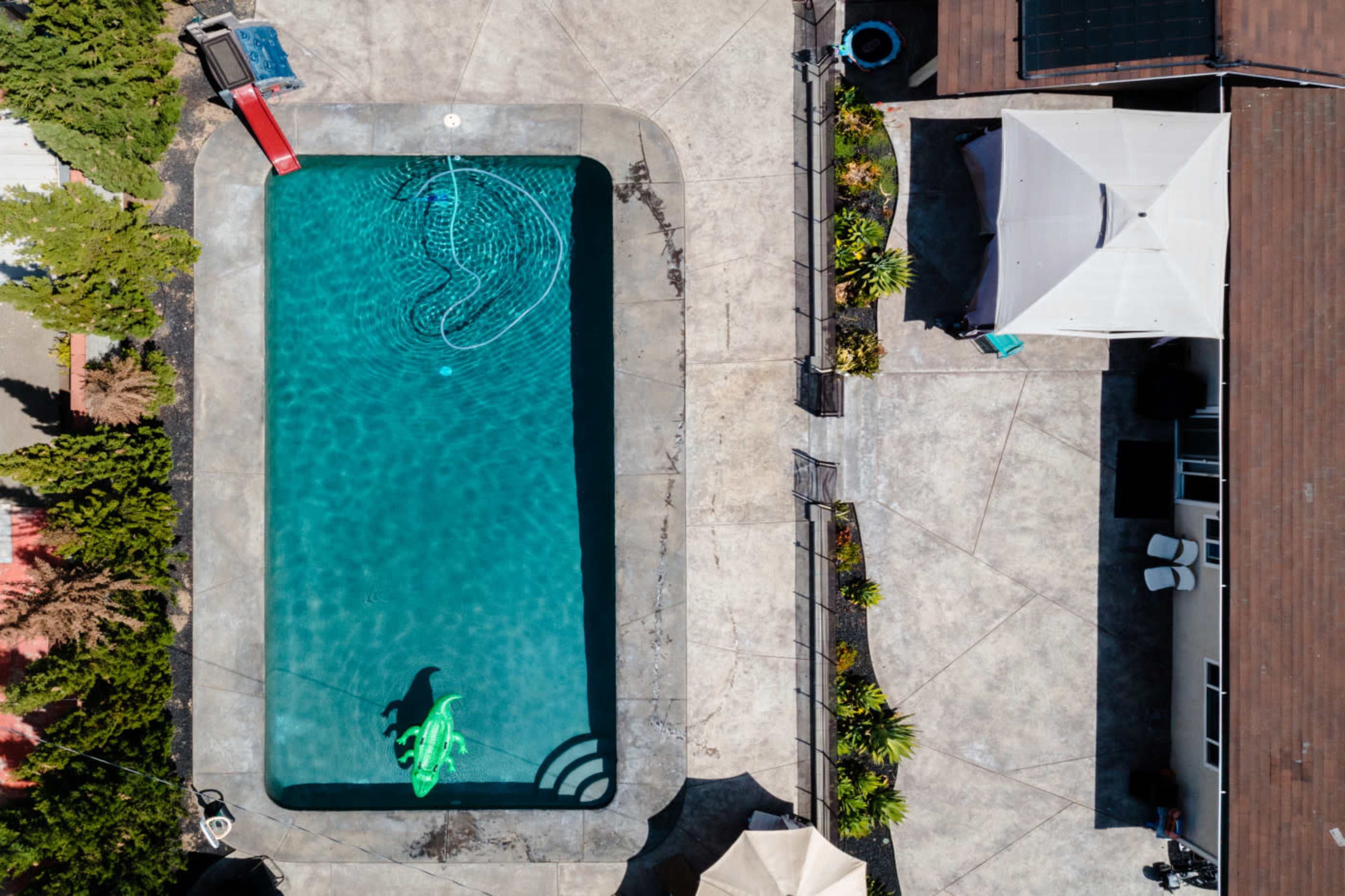 An aerial view of a rectangular swimming pool surrounded by concrete, featuring a green inflatable float and a shaded area with a gazebo.