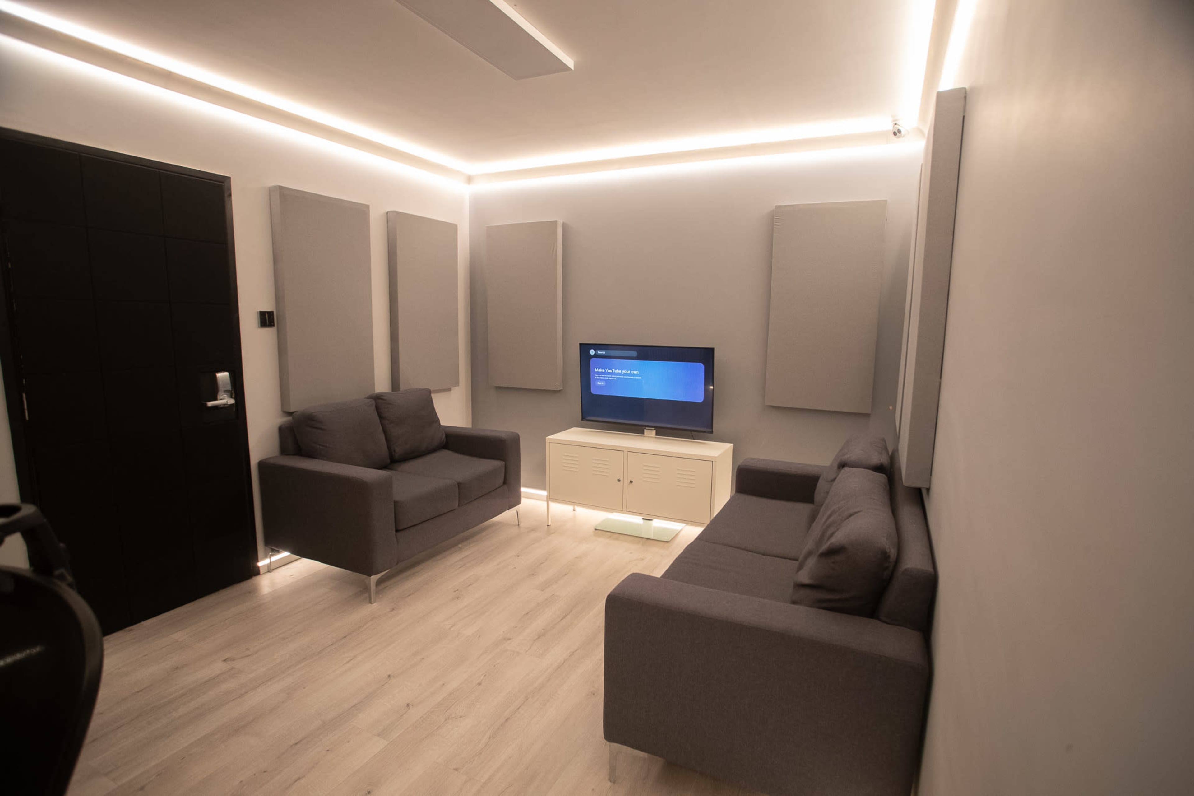 The image shows a modern room with two gray sofas facing a television, surrounded by sound-absorbing panels on the walls.