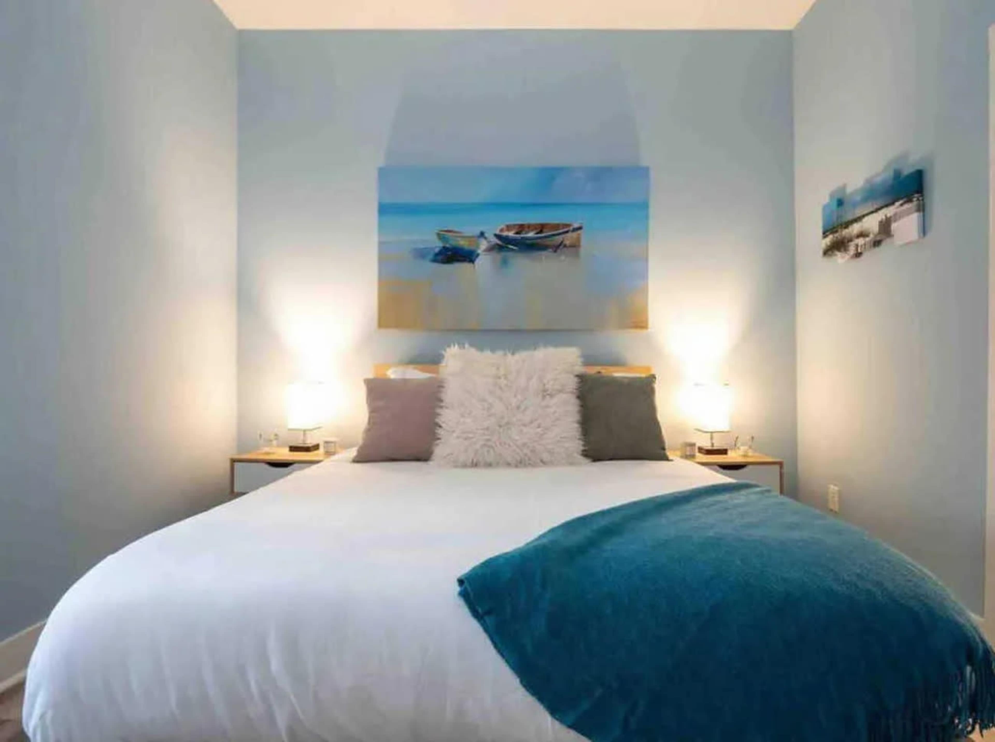 The image shows a neatly arranged bedroom with a double bed, light blue walls, and two bedside tables, each with a lamp, alongside decorative artwork depicting boats on the wall.