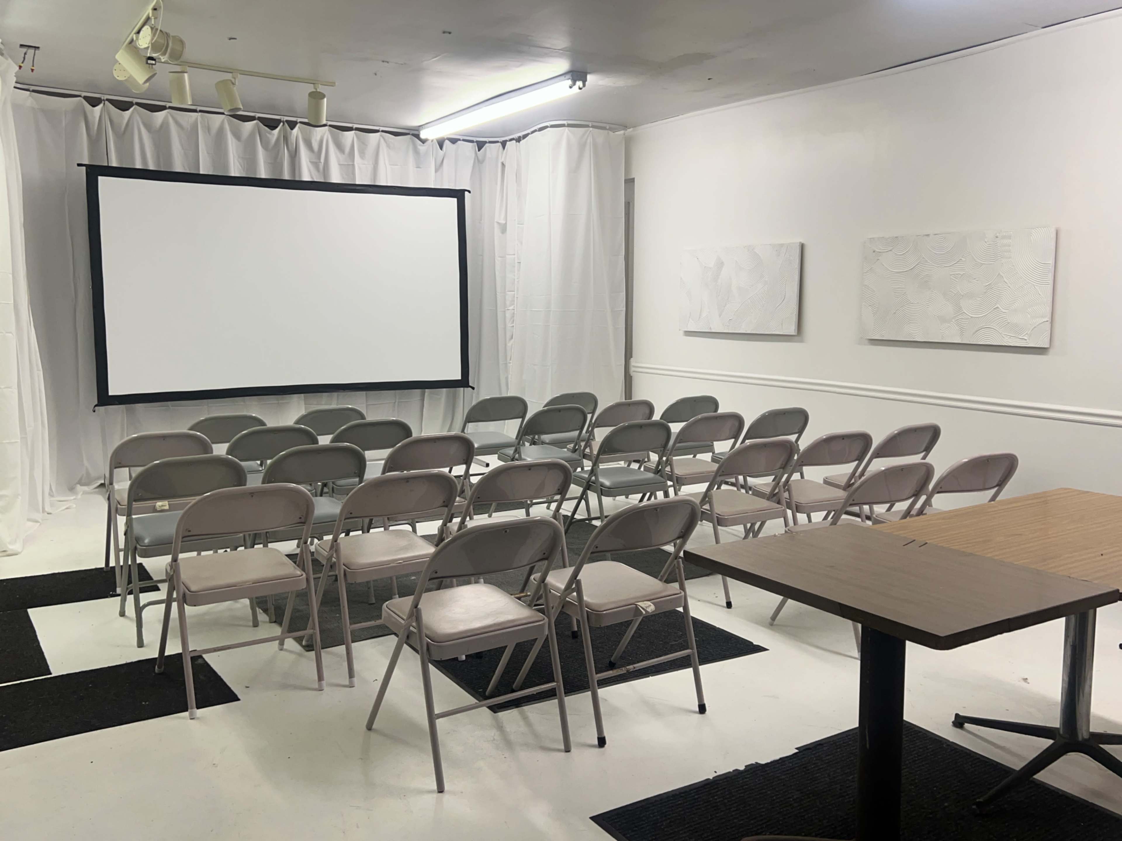 A small meeting room with rows of folding chairs facing a blank screen, a table to the side, and white drapes on the walls.