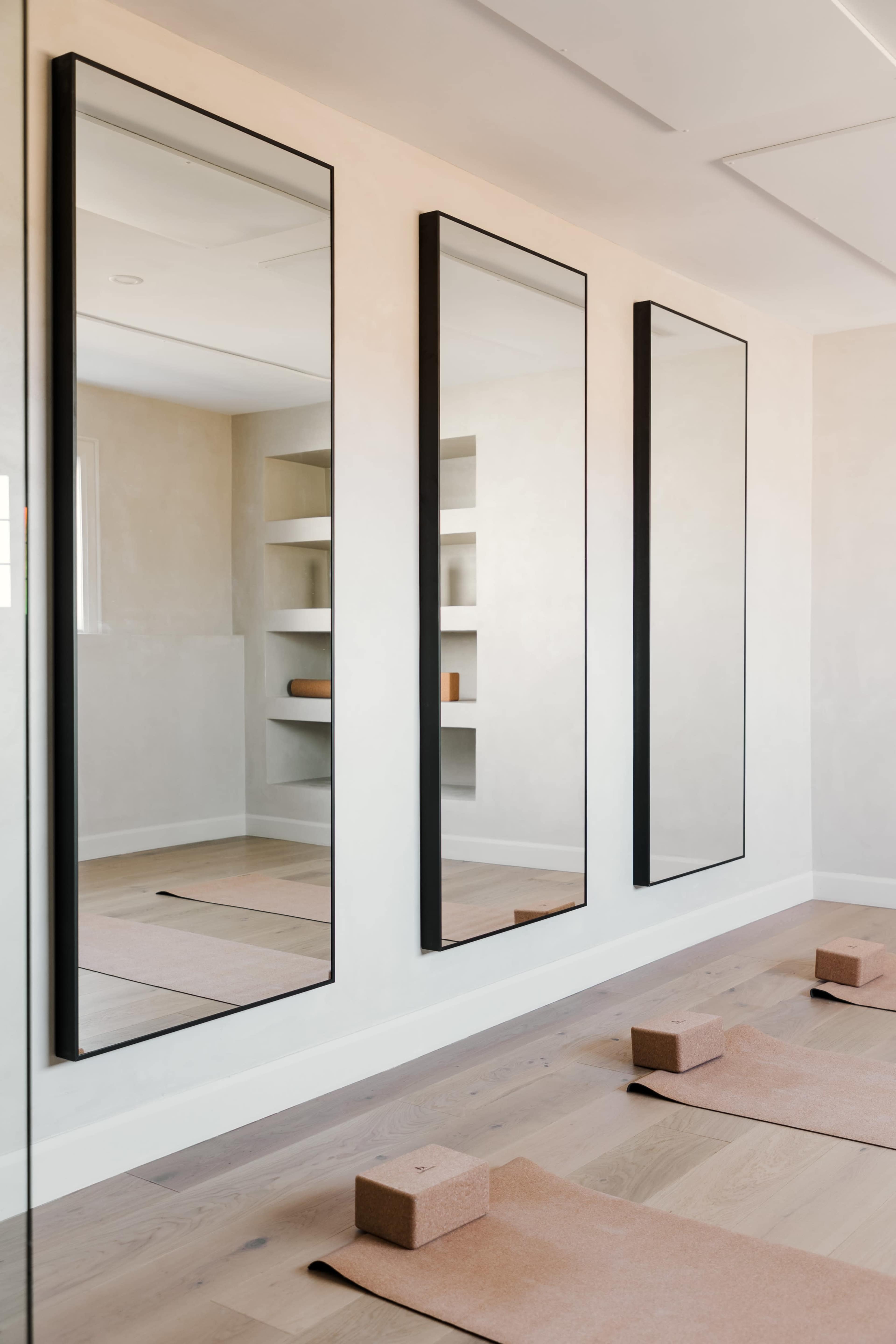 Three tall mirrors mounted on a light-colored wall, with a minimalist room containing shelves and yoga blocks on the floor.