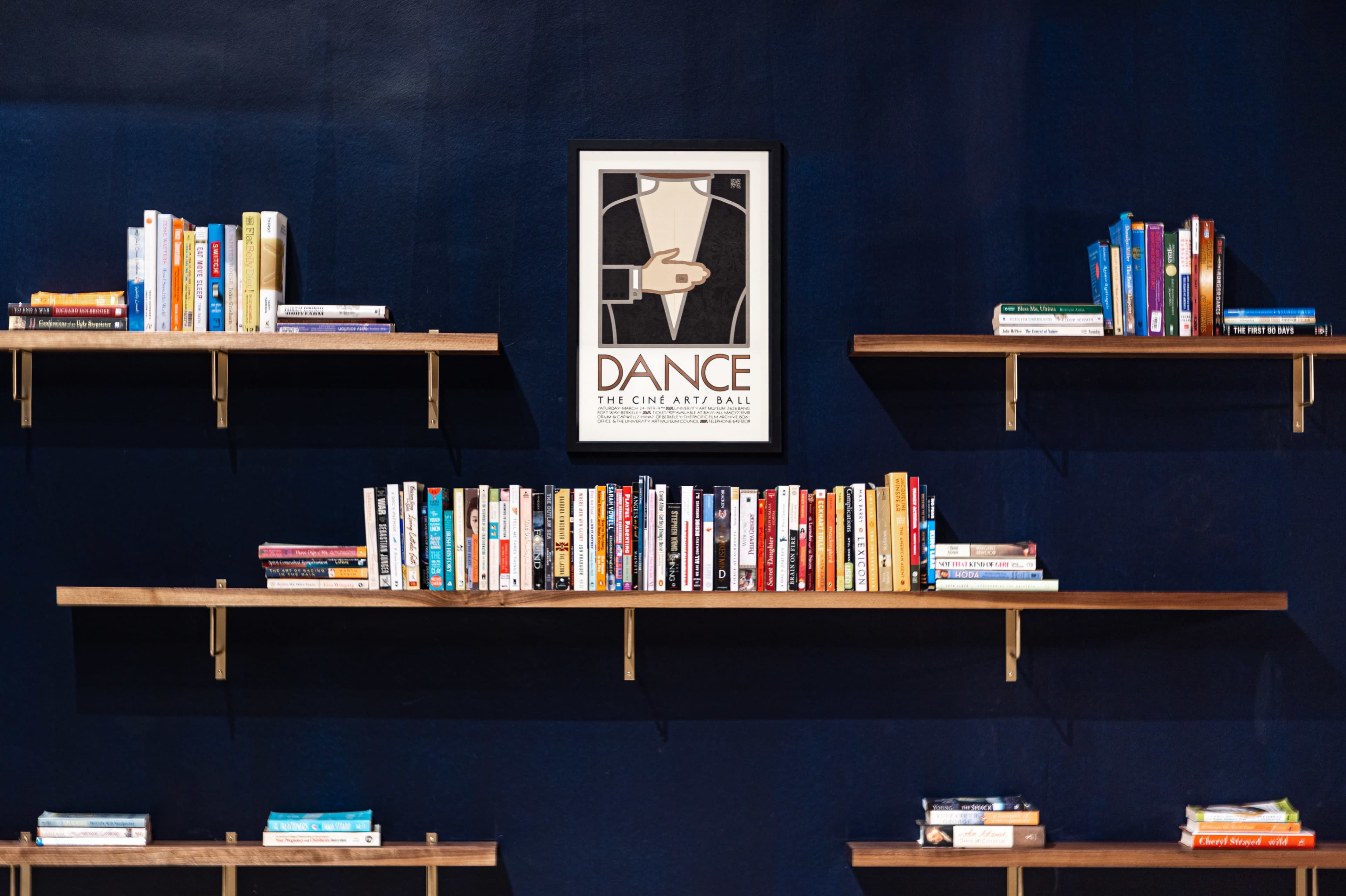 A dark blue wall featuring three wooden shelves filled with books and a framed poster titled "DANCE" hanging in the center.