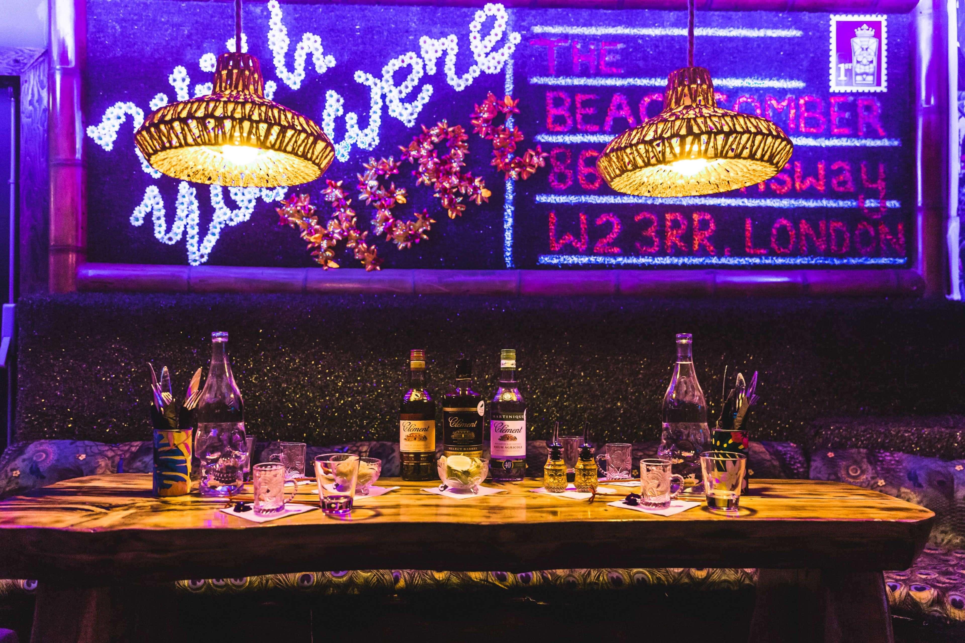 A wooden table is set with various bottles of alcohol and glasses, under two decorative lamps in a dimly lit bar adorned with colorful signage.