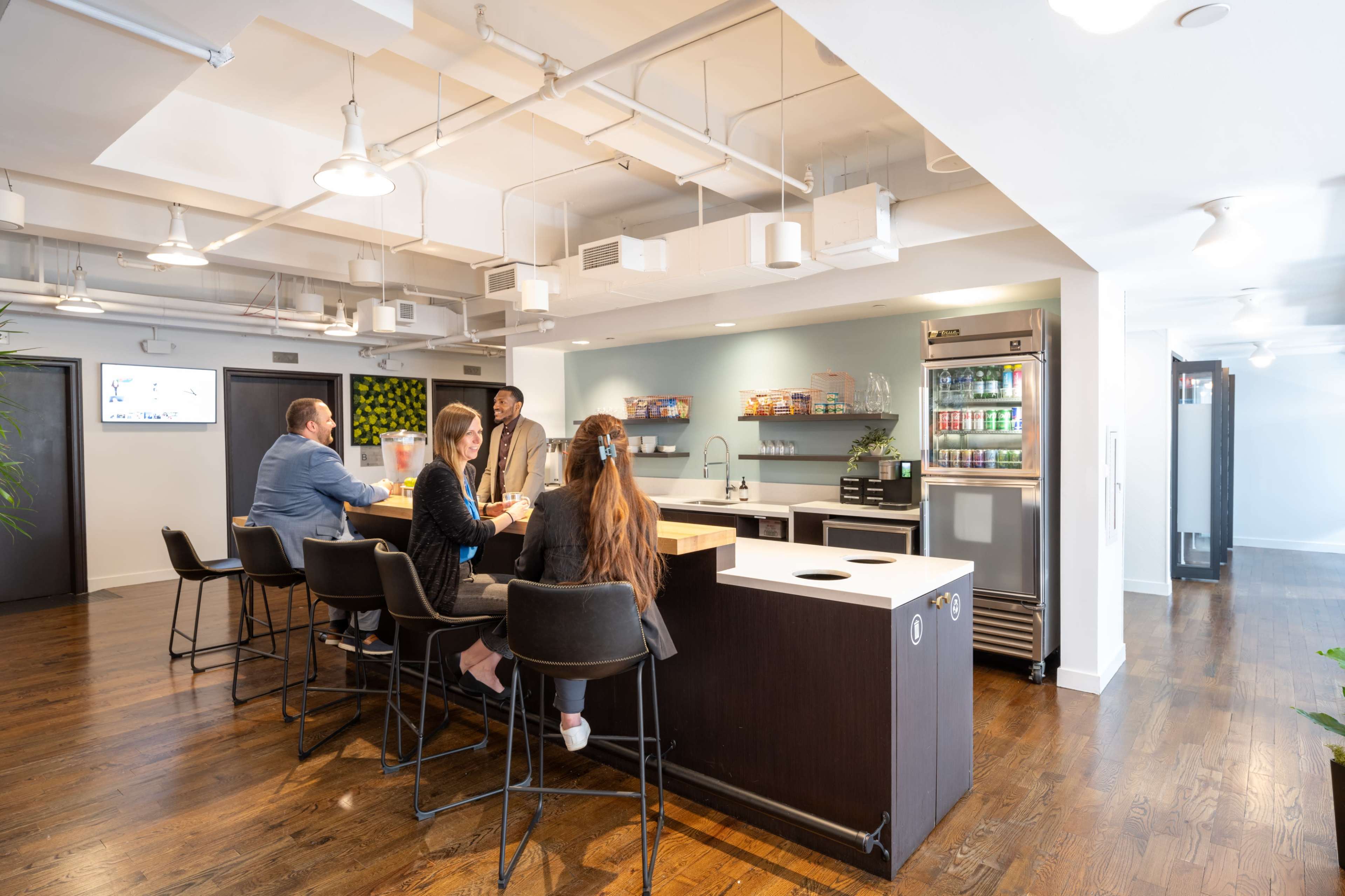 A group of four people is seated at a kitchen counter in a modern office space while a fifth person standing near them engages in conversation.