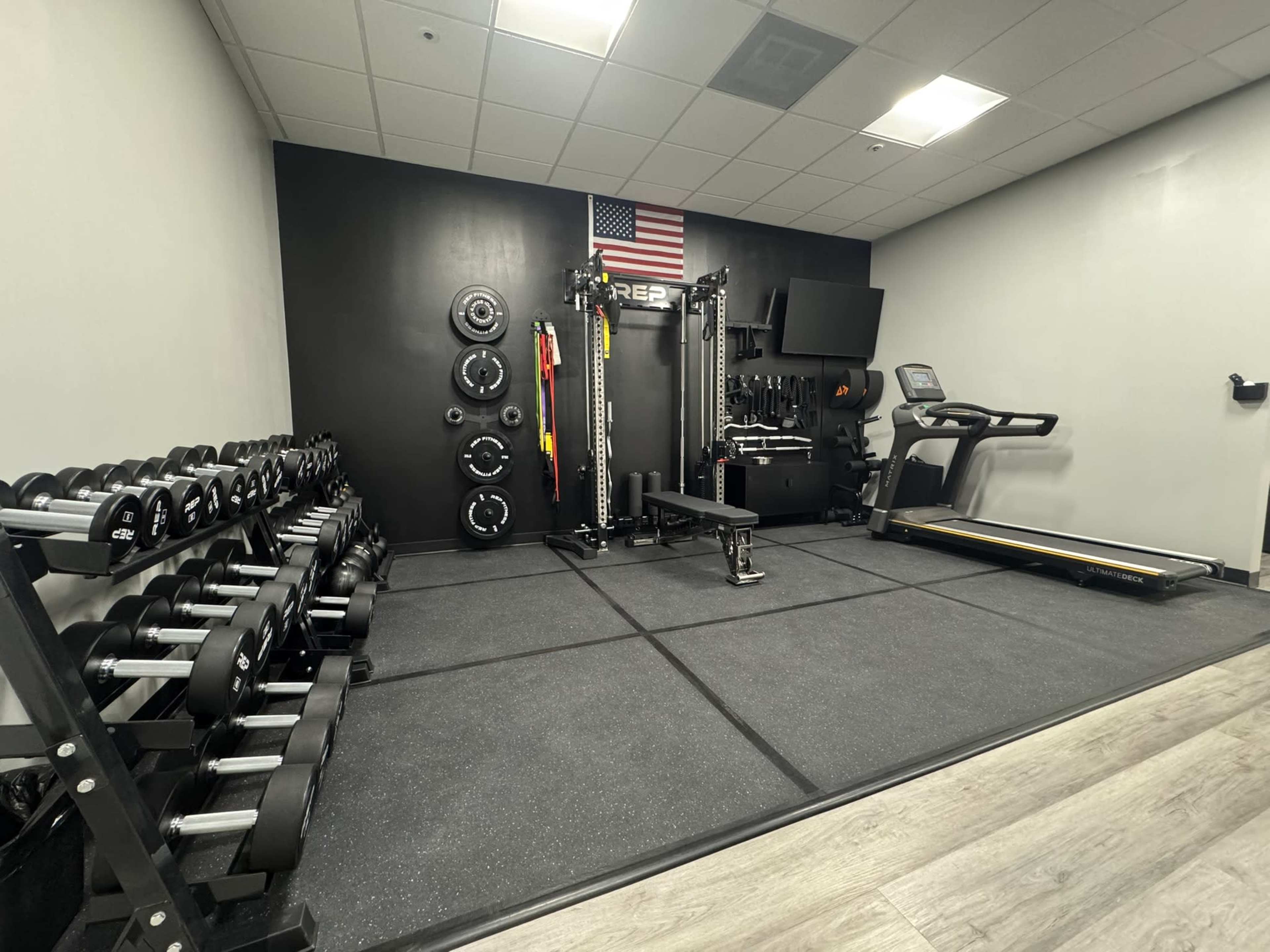 The image shows a well-equipped gym featuring a set of dumbbells, weight plates, a cable machine, a treadmill, and an American flag hanging on the wall.