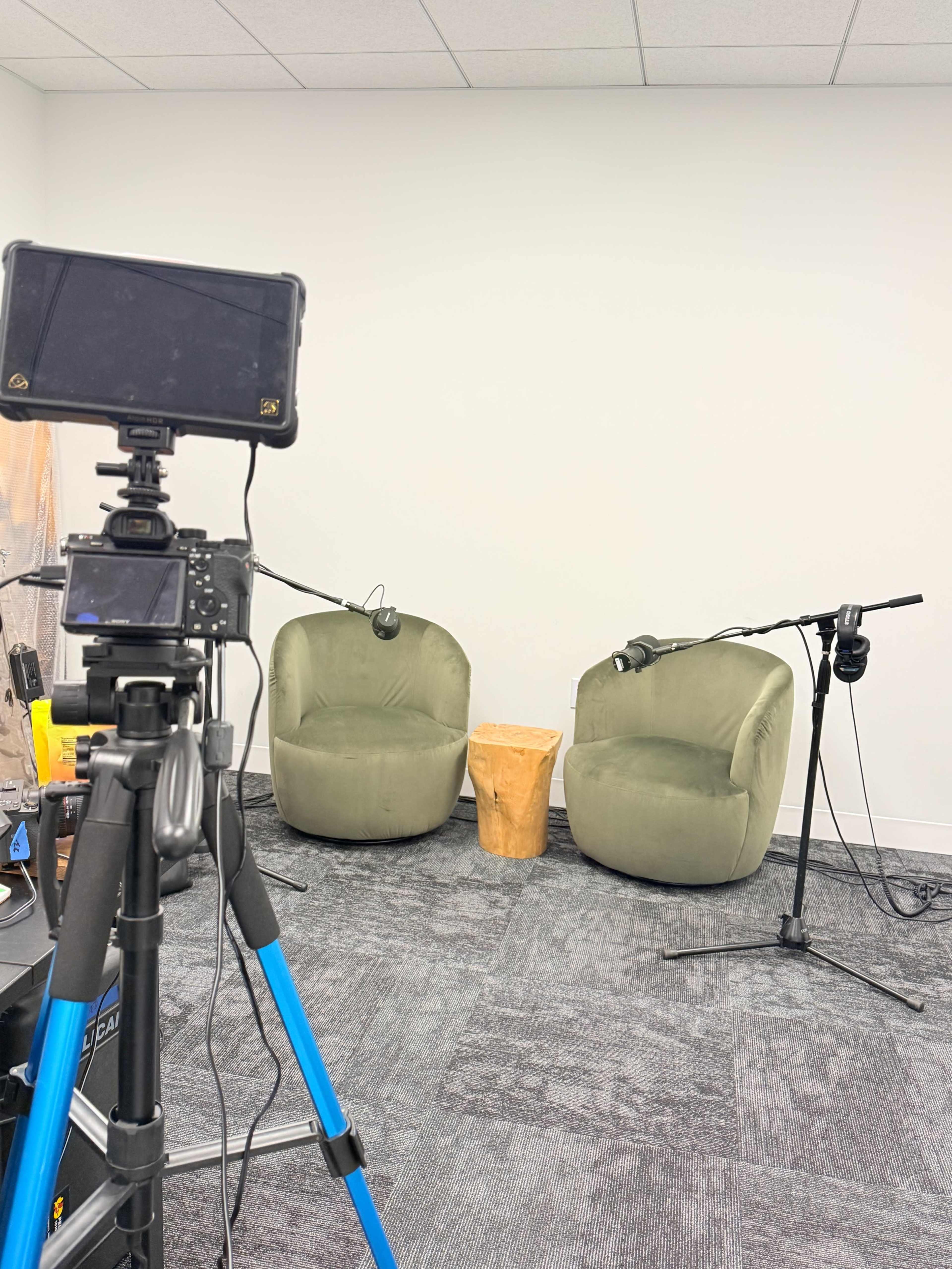 The image shows a podcast recording setup featuring two green chairs, a small wooden table, and a camera on a tripod pointing towards the chairs.