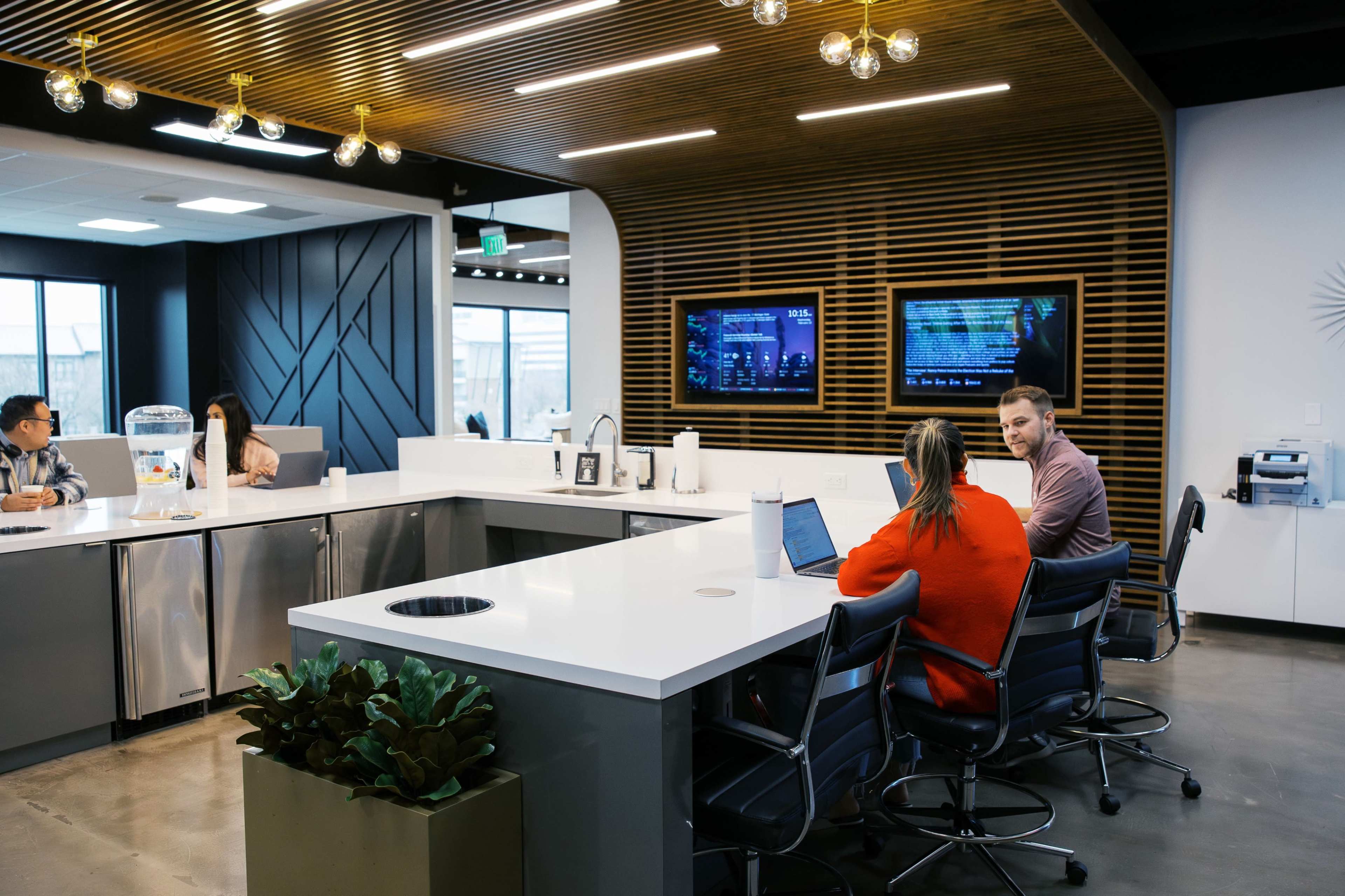 A modern office kitchen area features a large white countertop with seating, two people engaged in conversation, and screens displaying various data in the background.