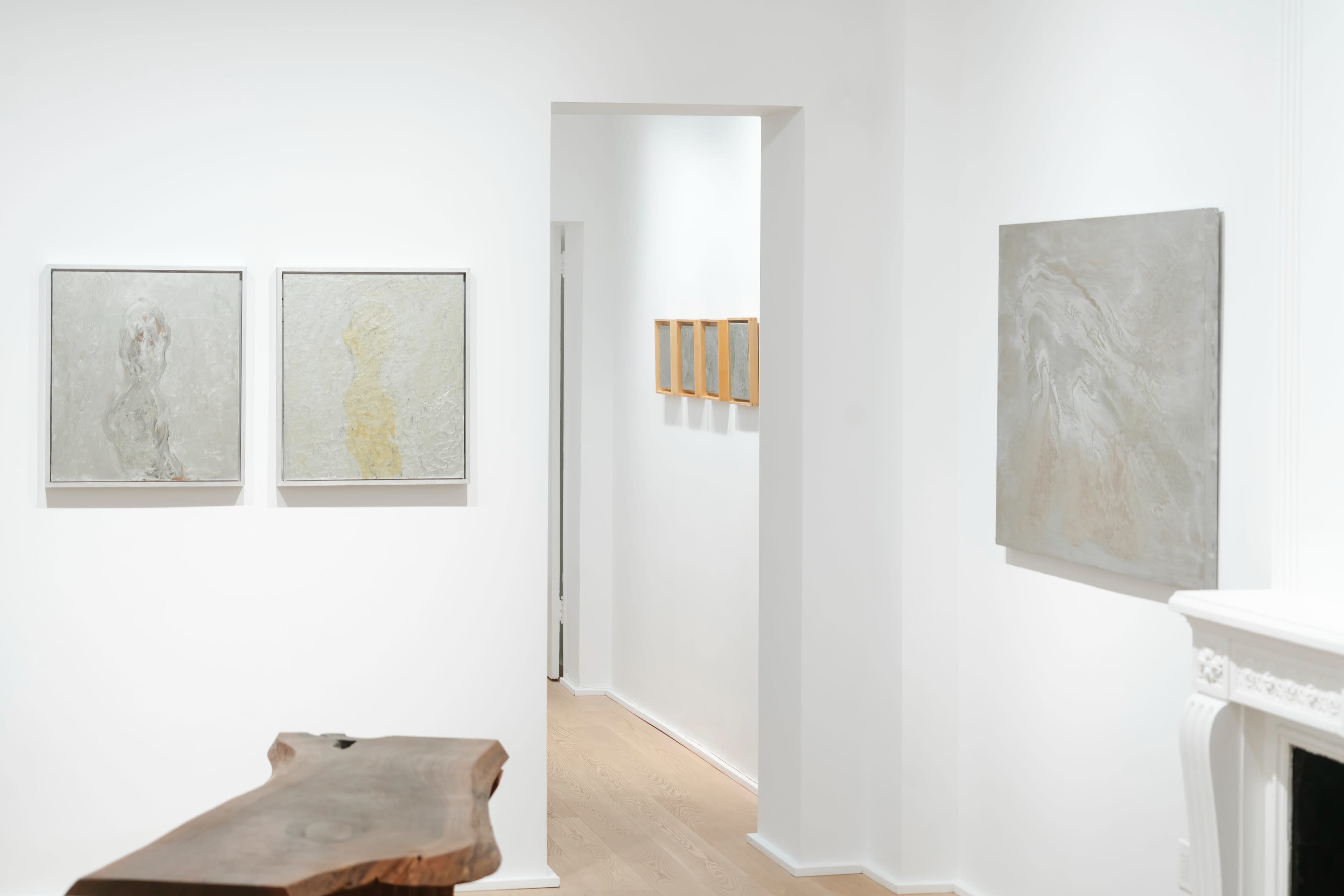 A minimalist gallery space featuring abstract artworks on white walls, with natural wood furniture and a corridor leading to additional framed pieces.