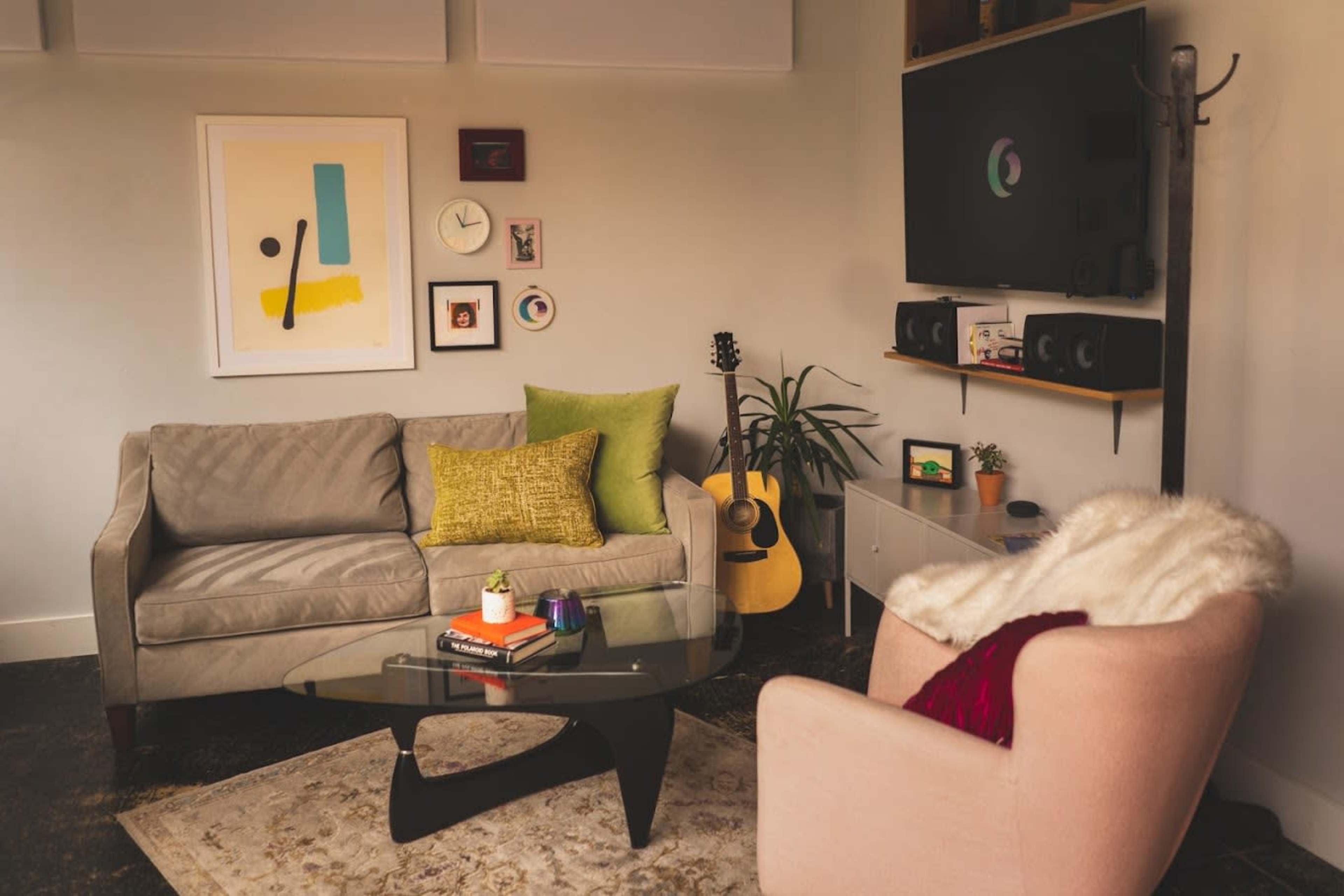 A cozy living room features a grey sofa, a pink armchair, a glass coffee table, and a guitar beside a wall-mounted TV.