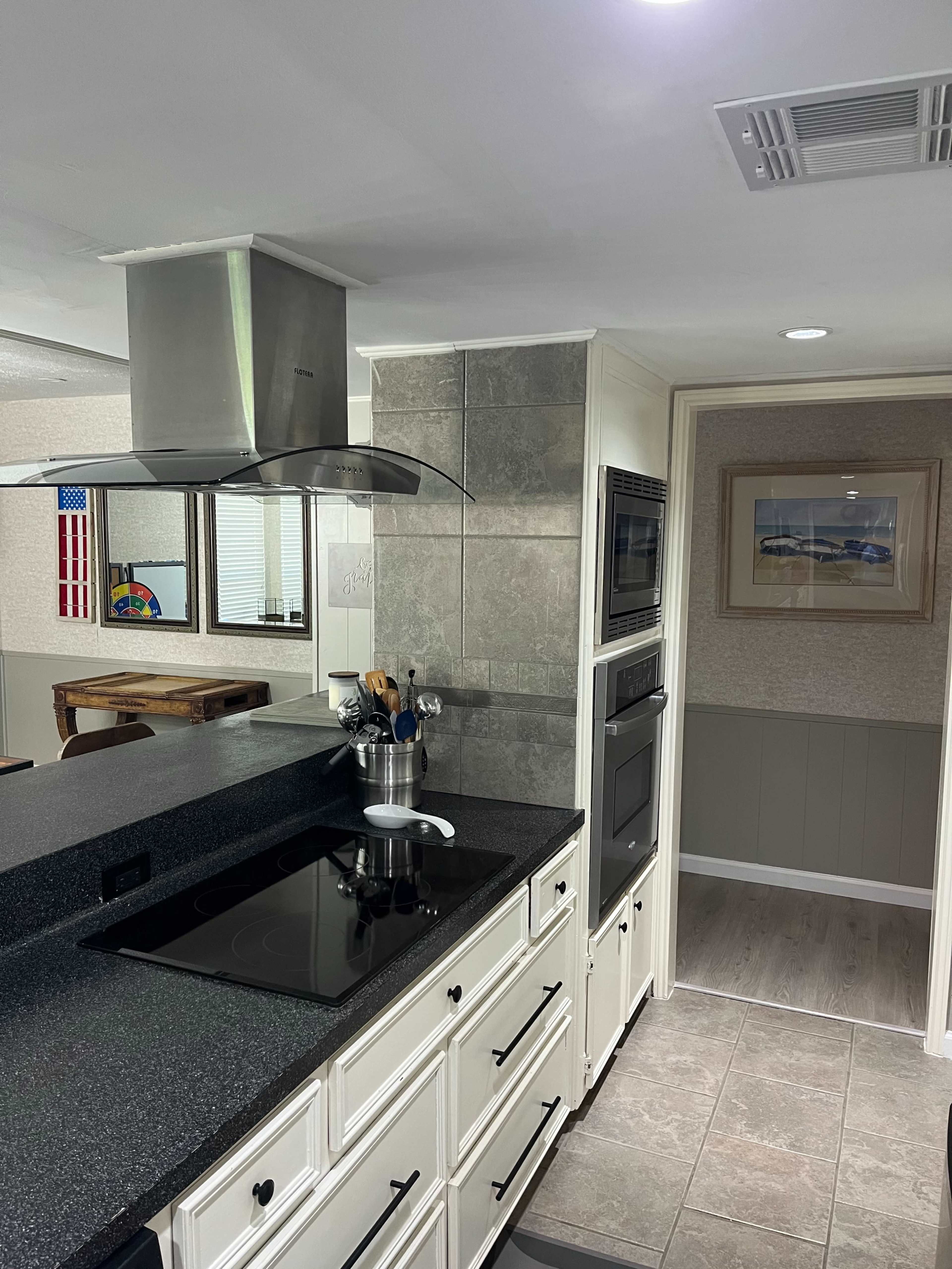 The image shows a modern kitchen with dark countertops, white cabinetry, and stainless steel appliances, leading to a hallway with framed artwork and a visible dining area.