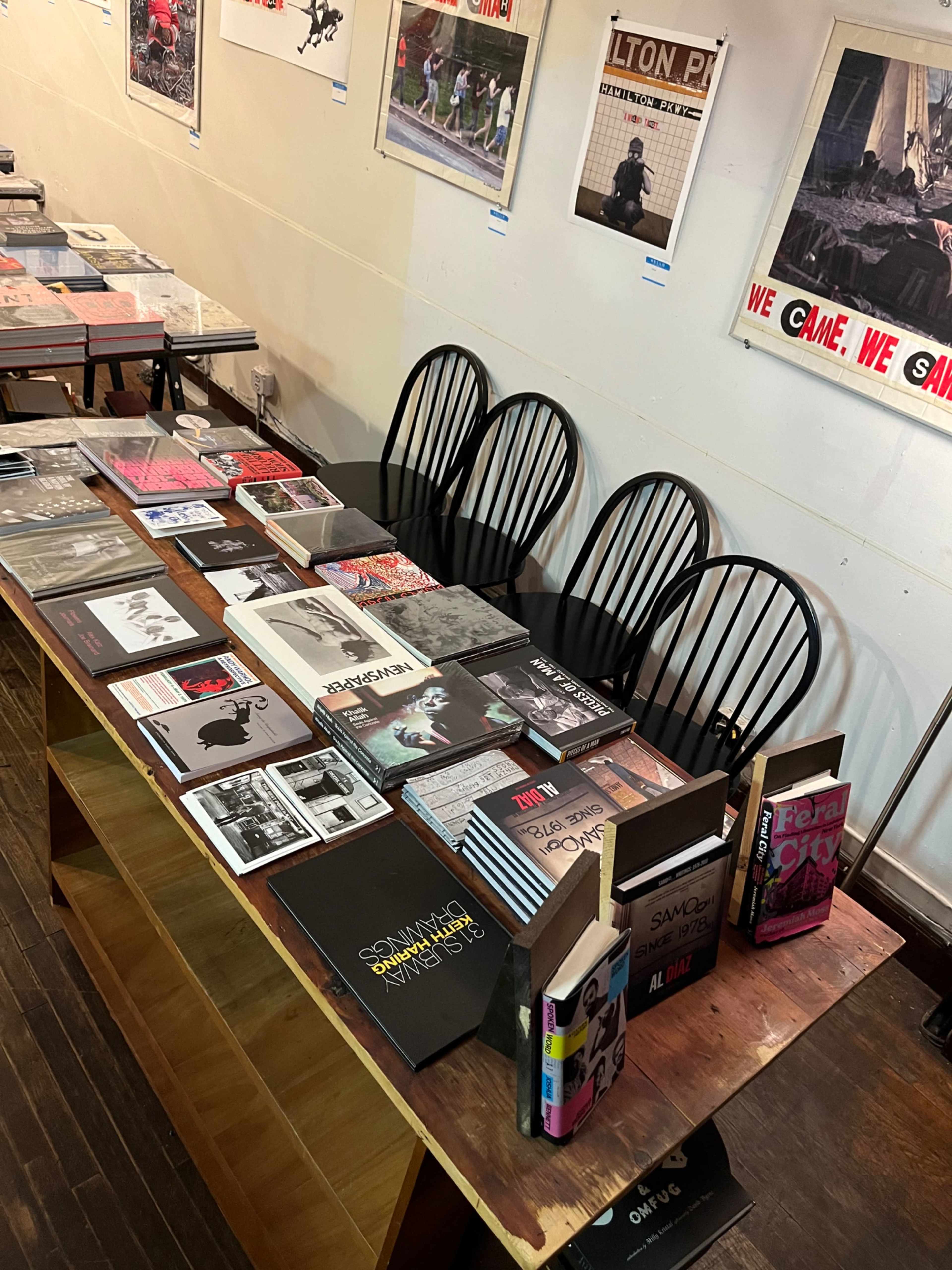 A long table displays various books and magazines, with several empty chairs arranged around it against a plain wall.