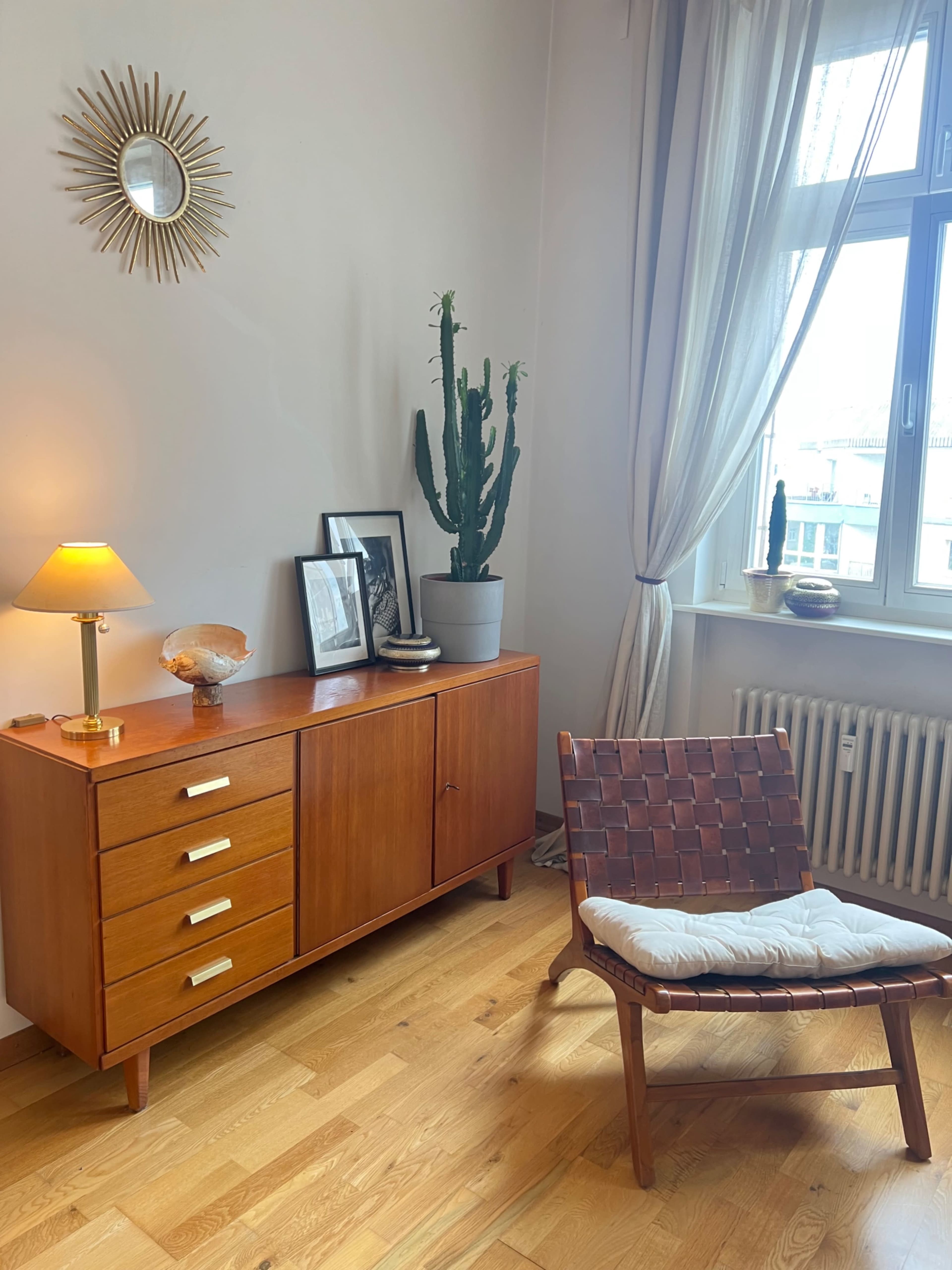 The image shows a cozy room featuring a wooden sideboard with drawers, a stylish chair, a potted cactus, and framed photographs on the wall, illuminated by a lamp.