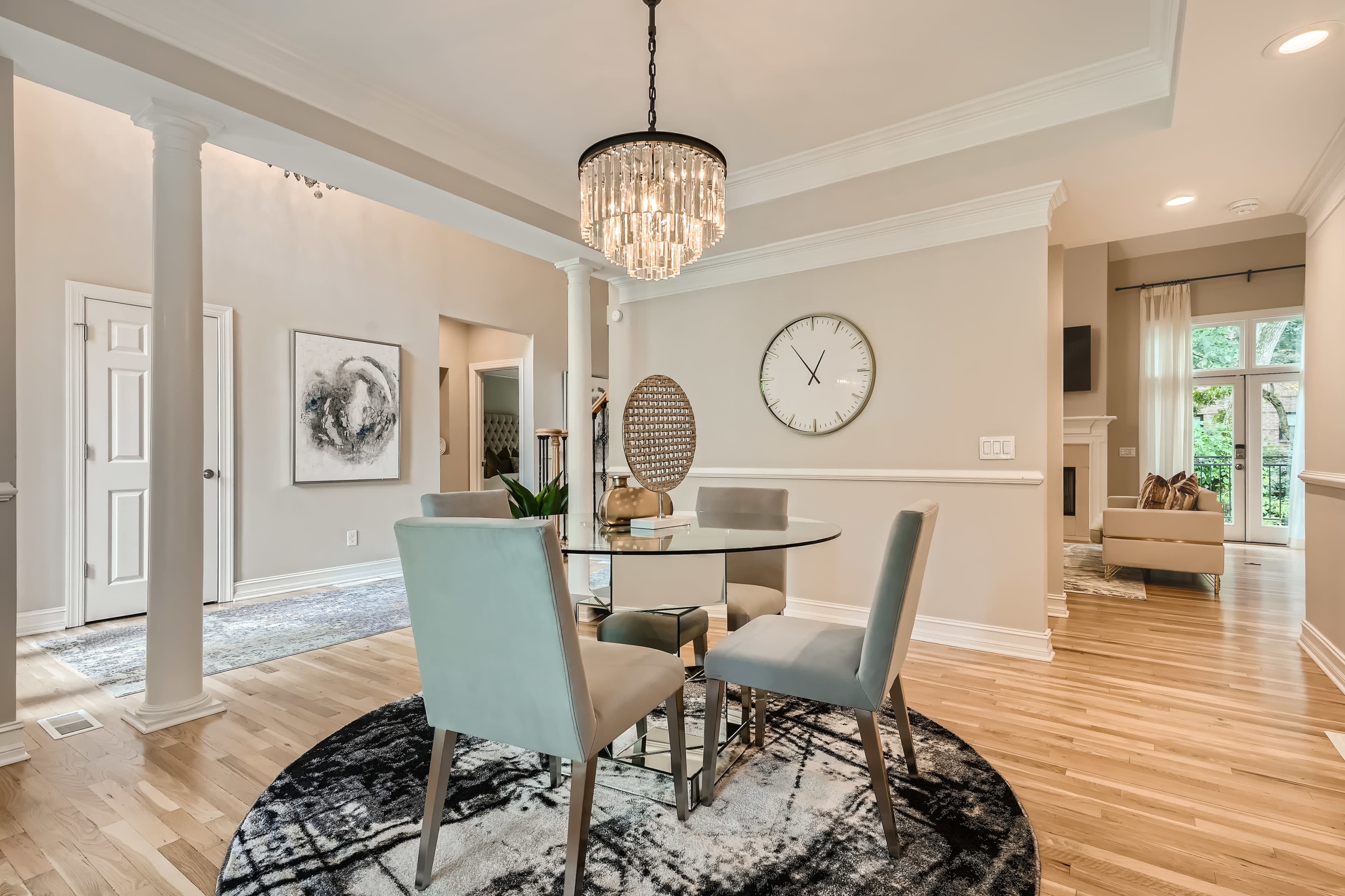 A dining area features a circular glass table surrounded by four light gray chairs, with a chandelier overhead and an oversized wall clock nearby.