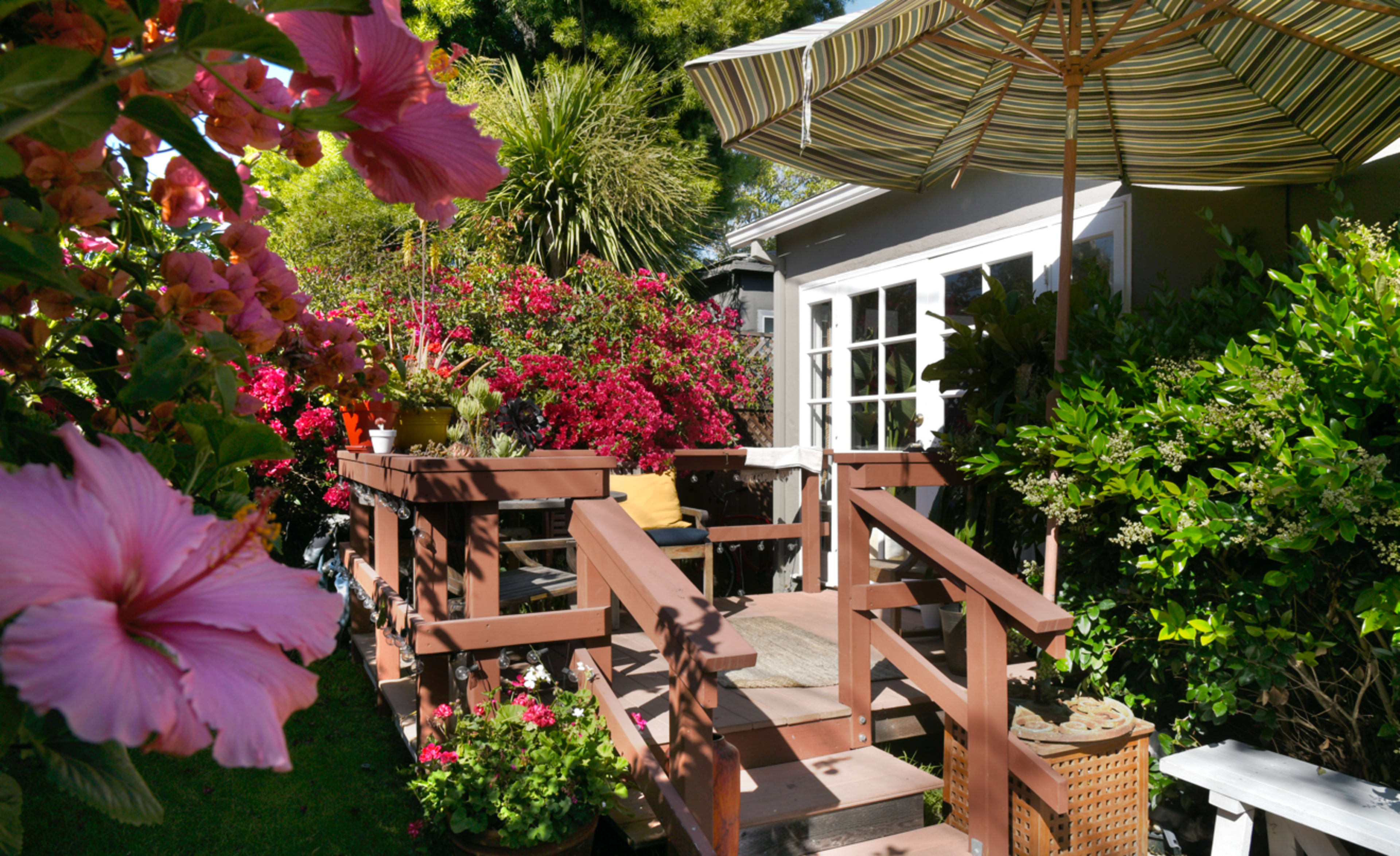 A wooden deck with a striped umbrella is surrounded by vibrant flowers and greenery near a house.