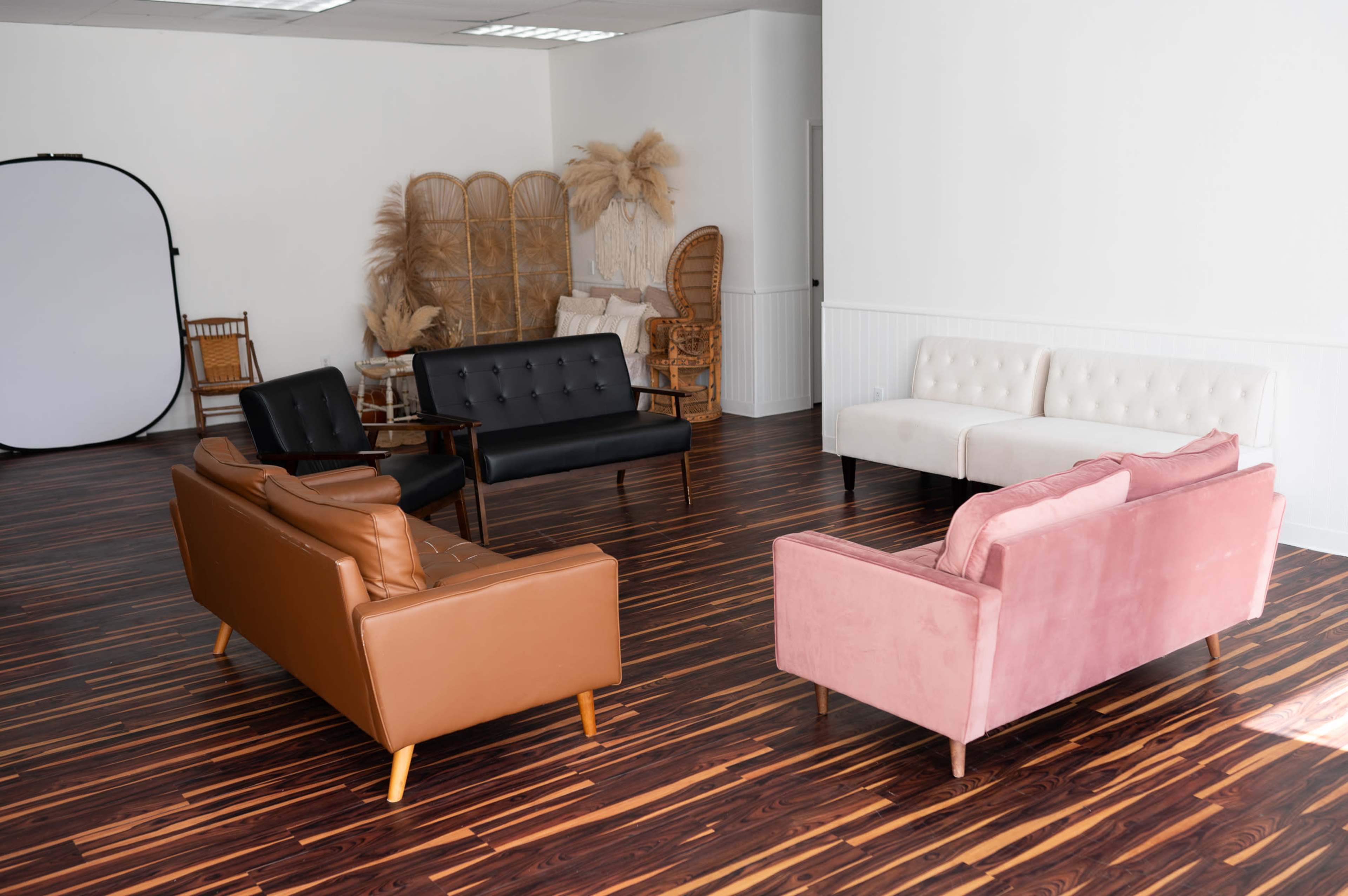 A bright, spacious room with wooden flooring, featuring four distinct sofas in different styles and colors, along with decorative elements like wicker furniture and plants.