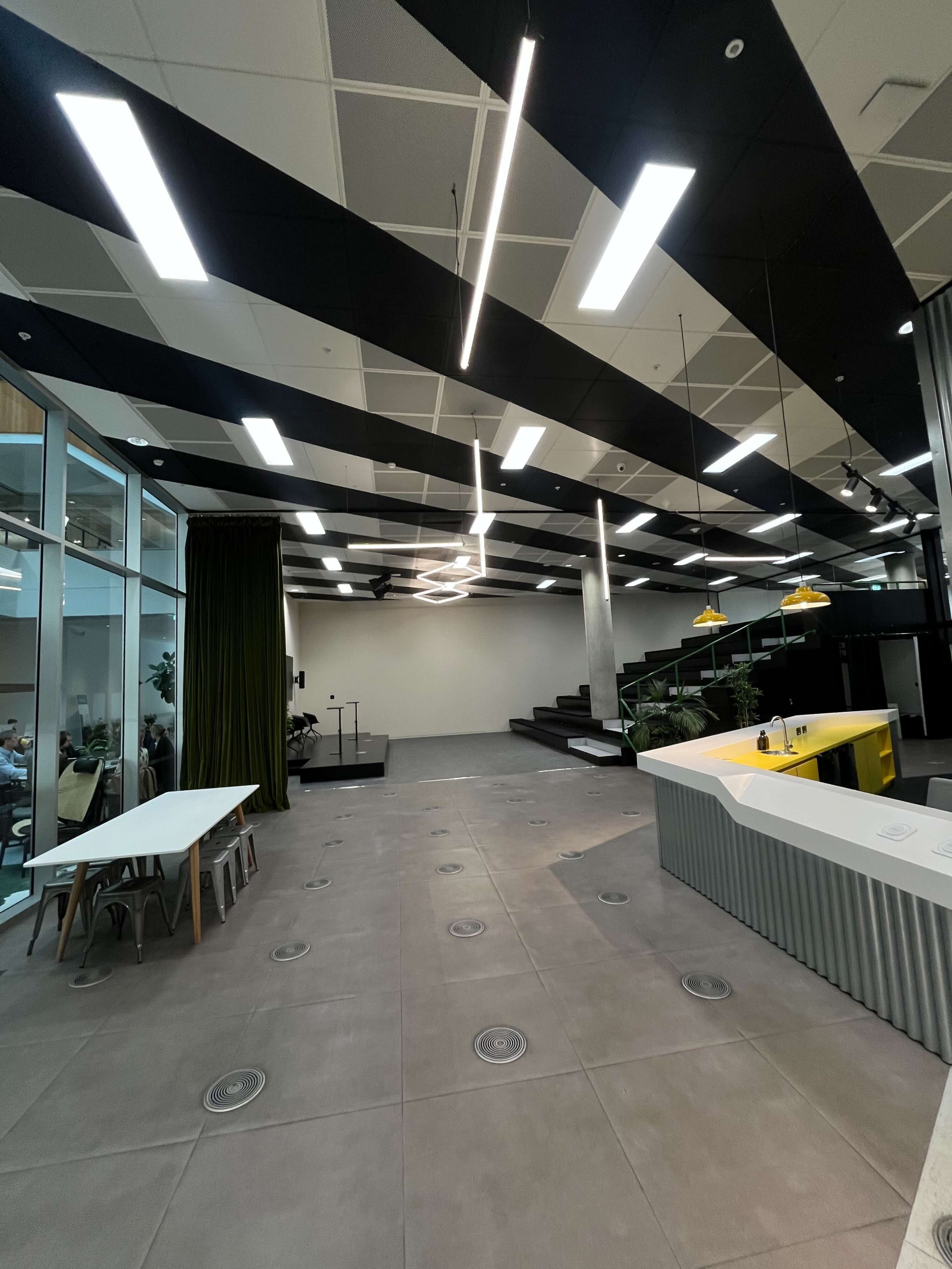 The image shows a modern office interior with a large open space featuring black and white ceiling lights, a kitchen area, and tiered seating in the background.